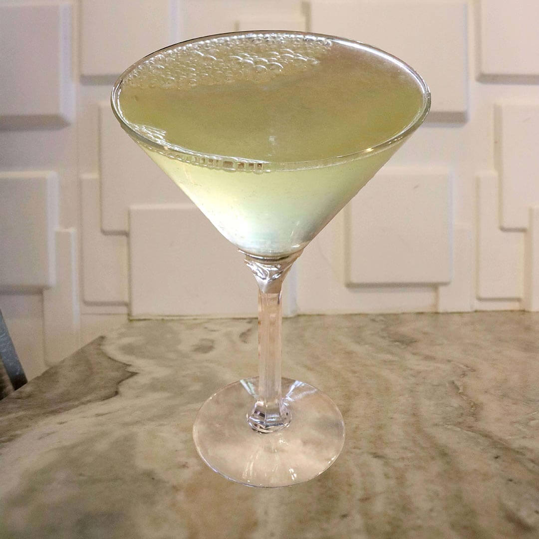 A pale, sparkling yellow cocktail served in a classic glass, featuring a cluster of effervescent bubbles along the rim.