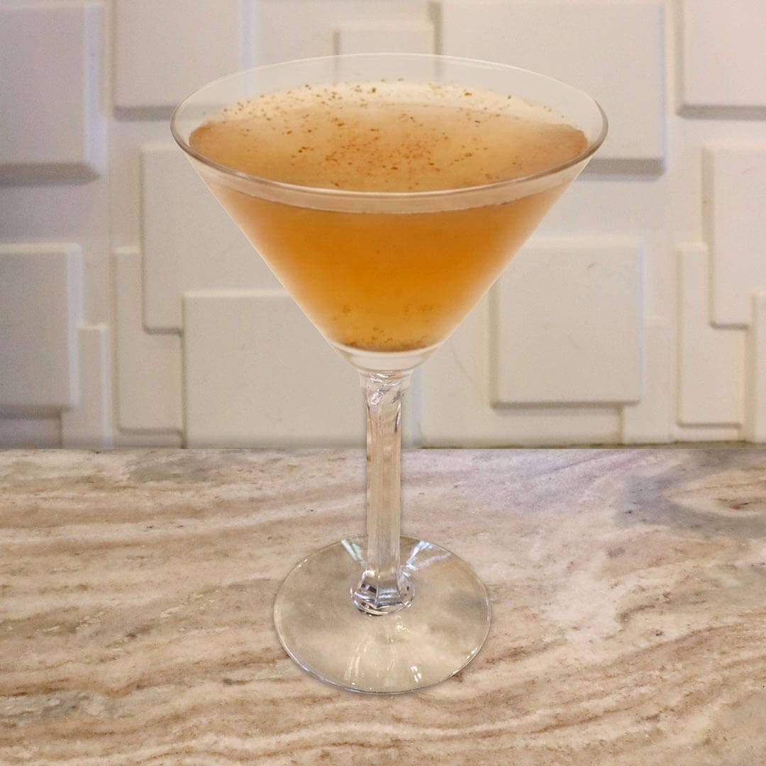 A golden, peach-colored cocktail served in a classic glass, featuring a light dusting of brown spices floating on the surface.