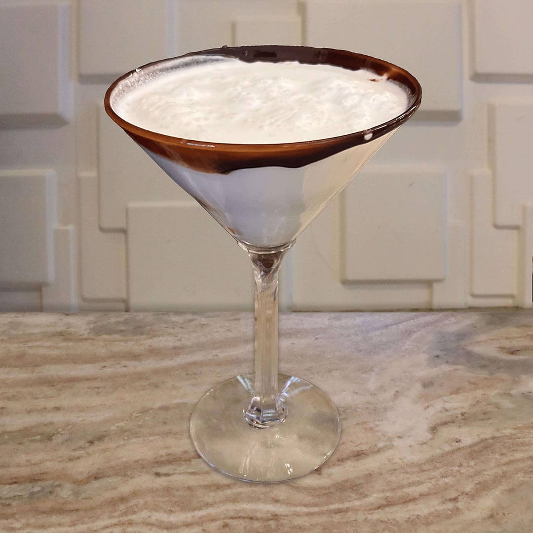 A creamy white cocktail with a thick layer of foam on top, served in a glass featuring a heavy ring of chocolate syrup around the rim.