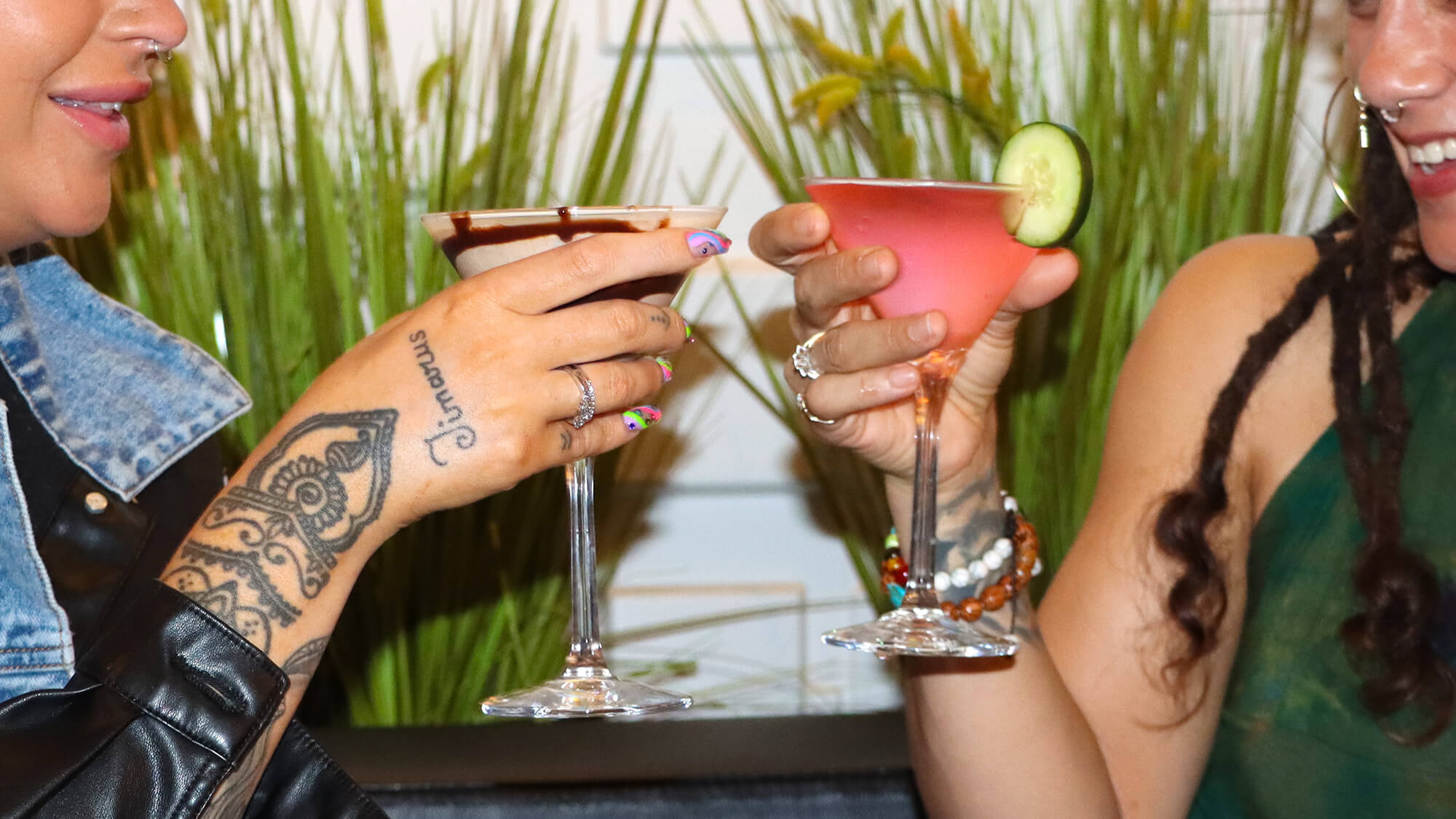 Two smiling women clinking a chocolate martini and a pink fruit martini with a cucumber garnish in front of decorative green grass.
