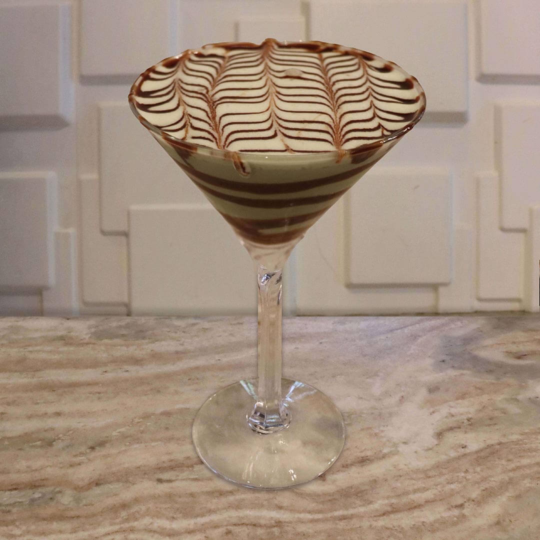 A creamy, pale mint-green cocktail featuring thick chocolate swirls inside the glass and chocolate feathering on top.