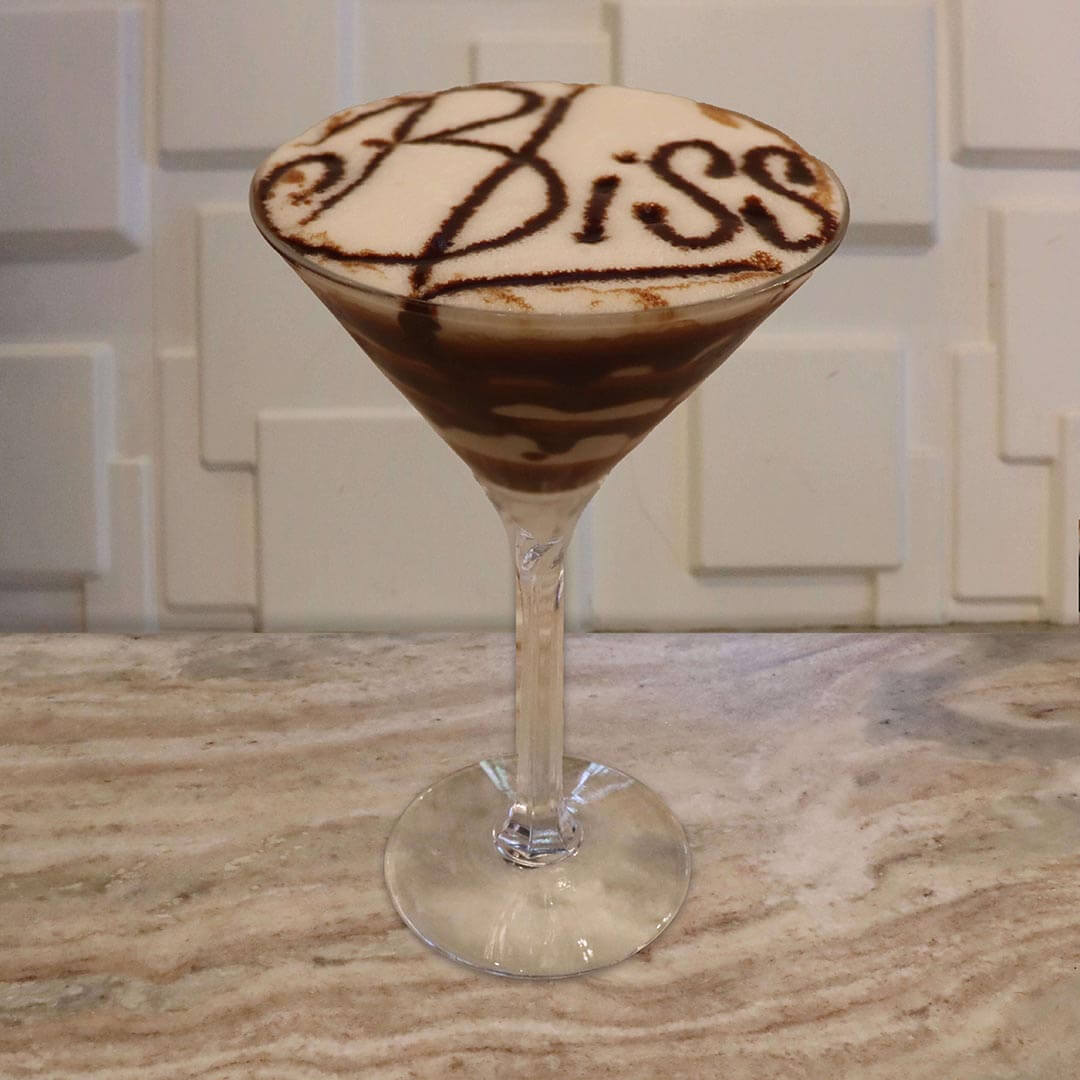 A creamy, pale tan cocktail featuring delicate chocolate syrup swirls coating the inside of the glass.