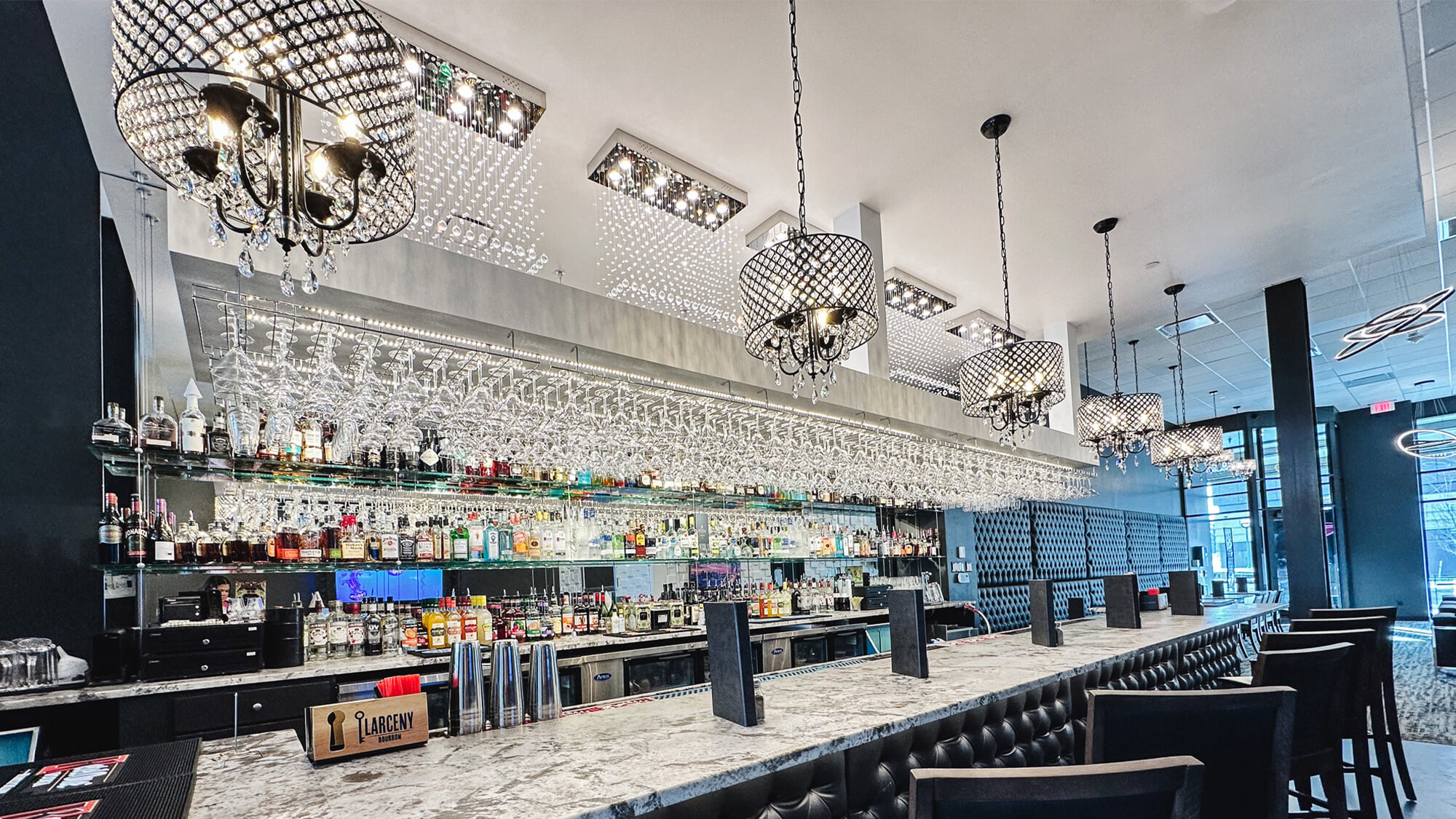 Expansive marble bar at The Stuffed Olive in Omaha featuring tufted black leather, tall barstools, crystal chandeliers, and a massive illuminated liquor display with hanging glassware.
