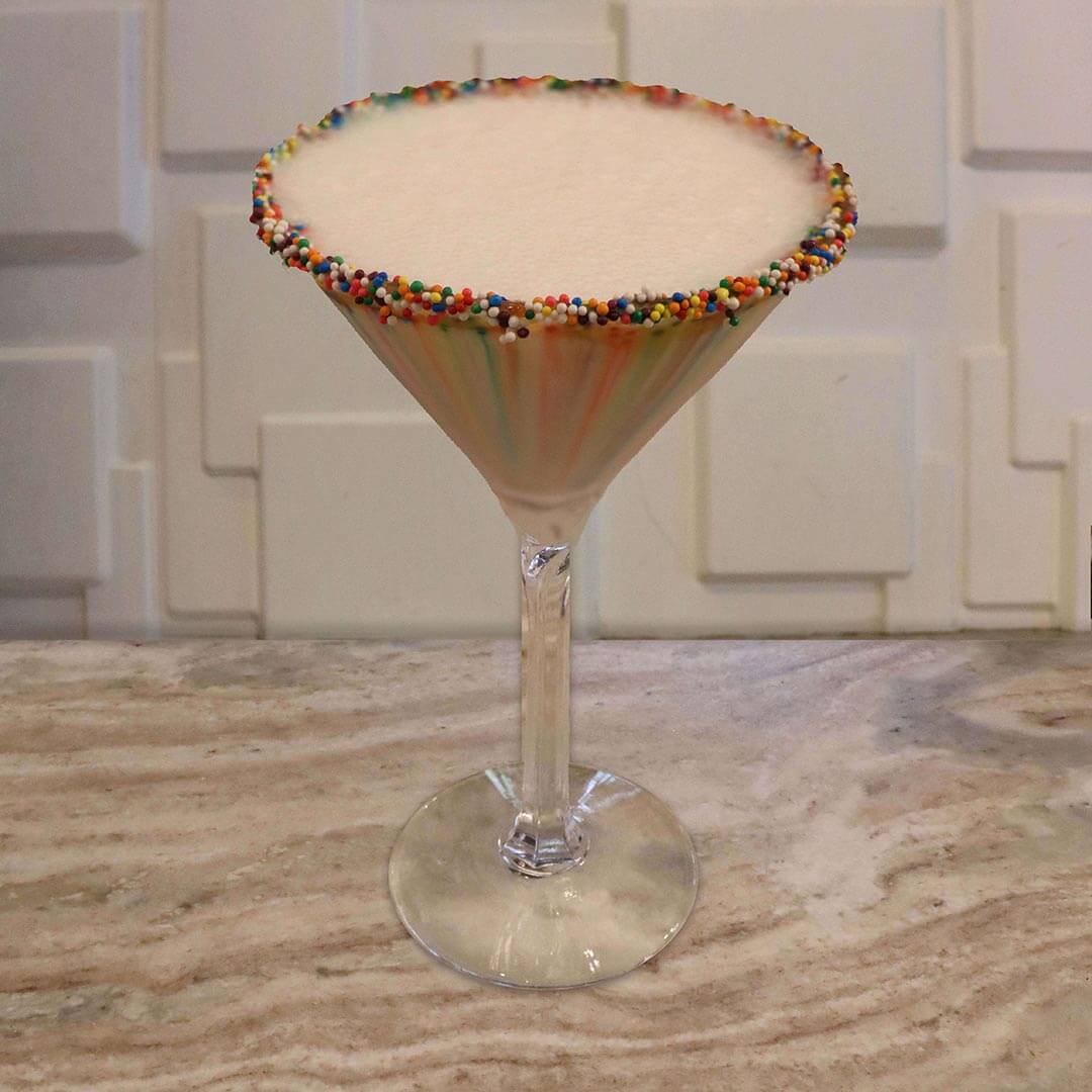 A creamy, opaque white cocktail featuring a brightly colored rainbow sprinkle rim.