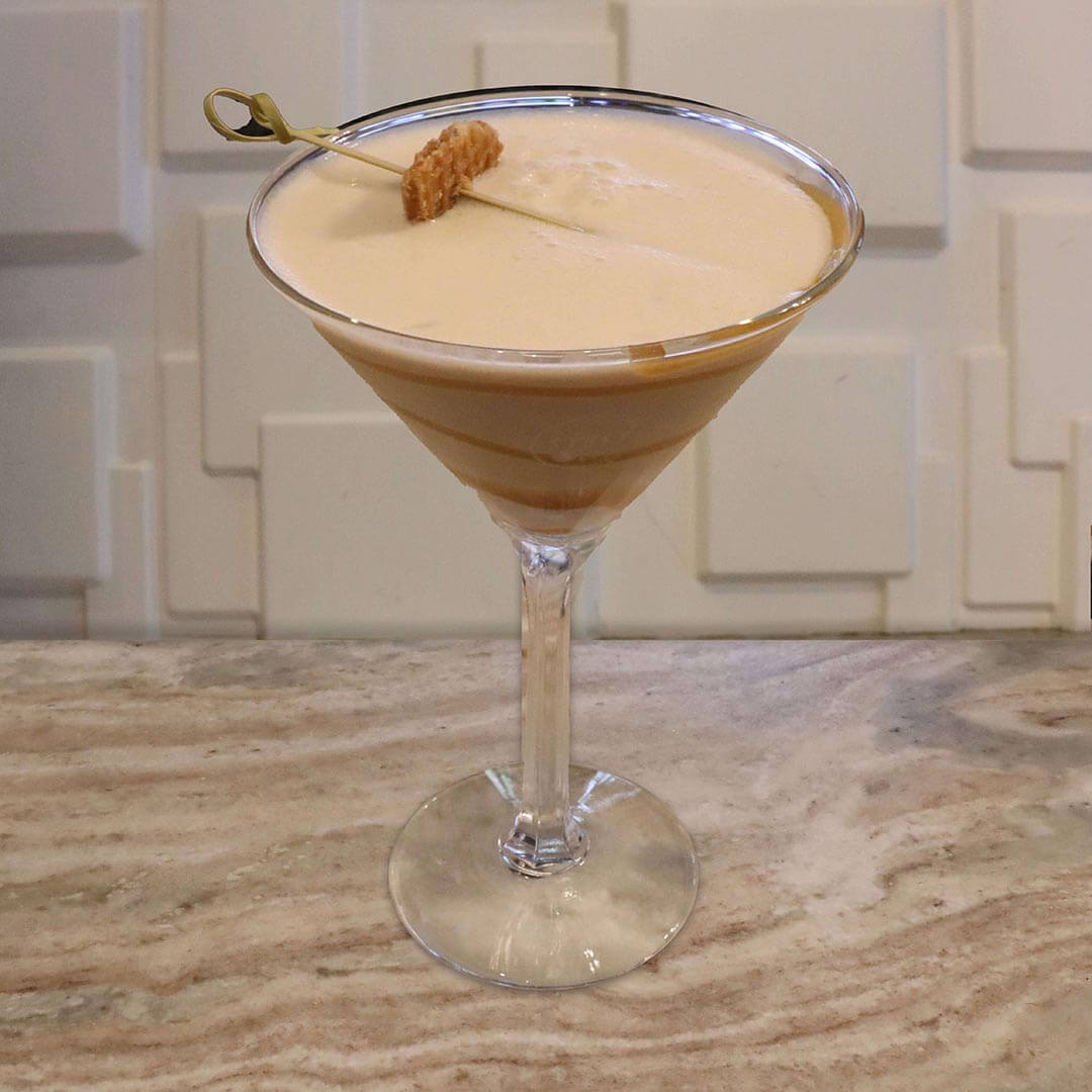 A creamy, light tan cocktail featuring thick caramel swirls inside the glass, garnished with a mini caramel waffle cookie.