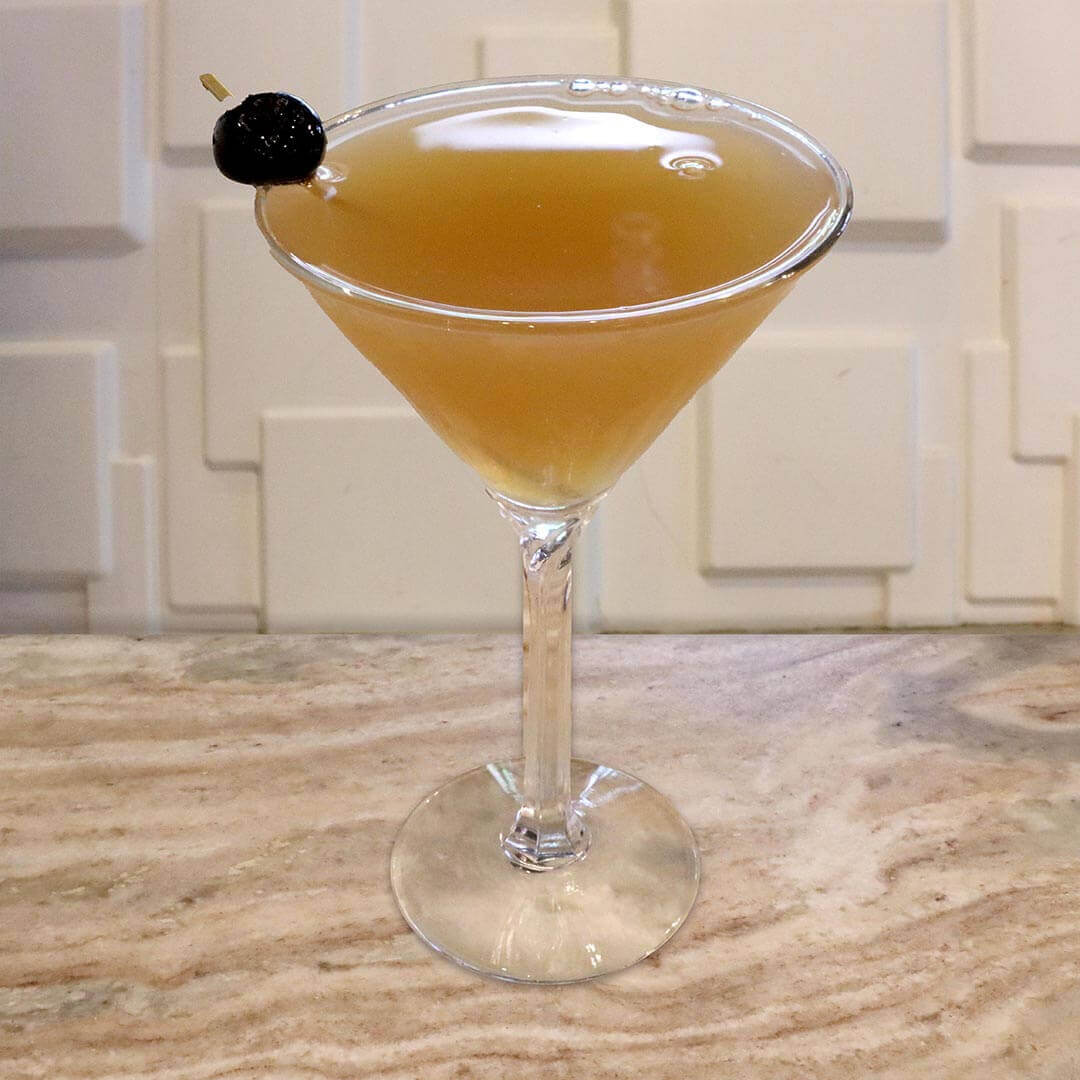 A light golden-amber cocktail in a chilled glass, garnished with a dark cherry.