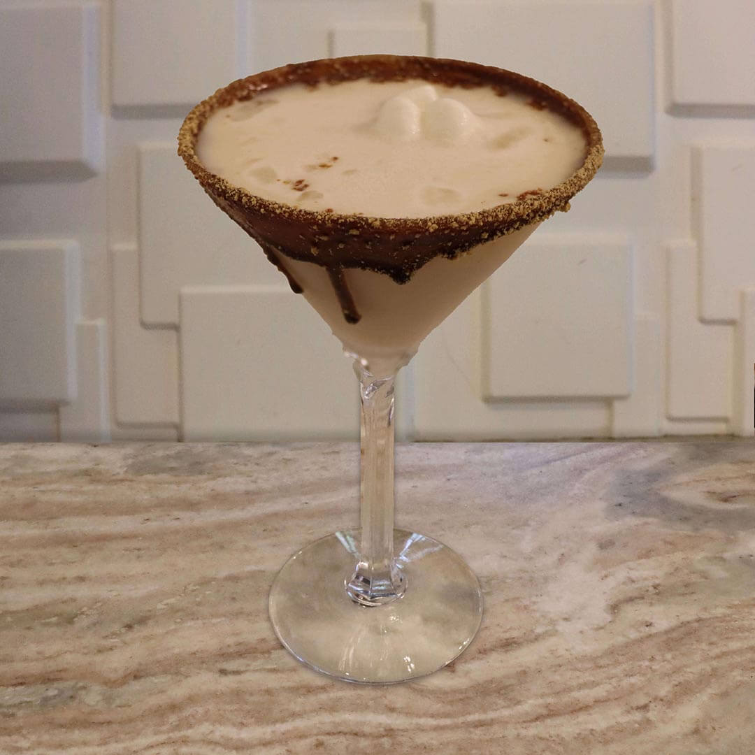 A creamy, light brown cocktail featuring a chocolate graham cracker rim and floating miniature white marshmallows.