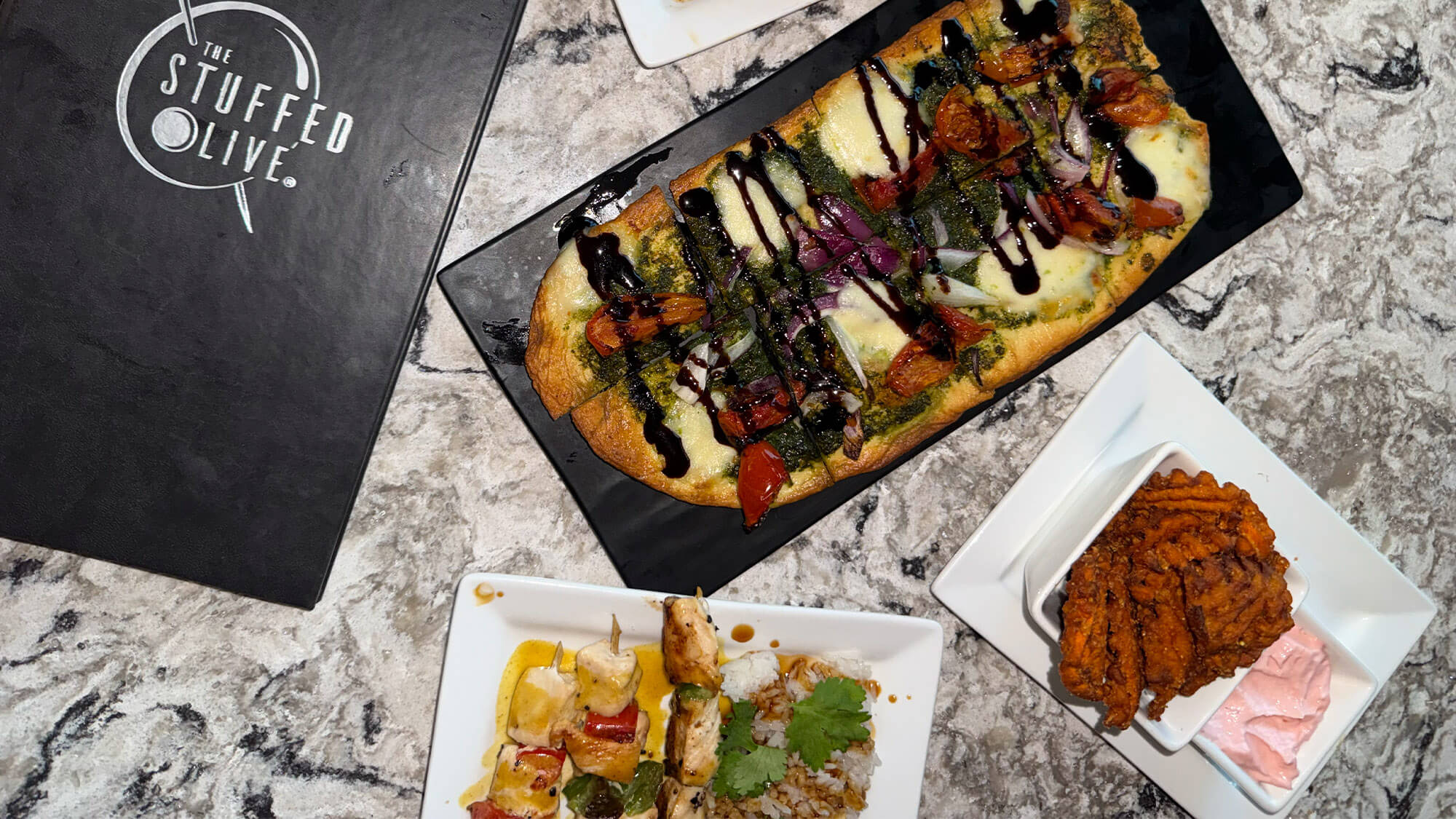 Overhead view of a marble table displaying The Stuffed Olive menu alongside a pesto and balsamic flatbread, chicken skewers over rice, and sweet potato fries with pink dipping sauce.