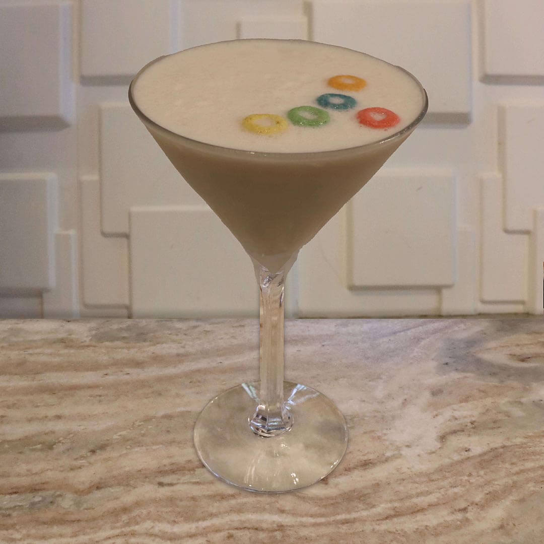 A creamy, pale pinkish-white cocktail garnished with a colorful floating ring of fruit cereal pieces.