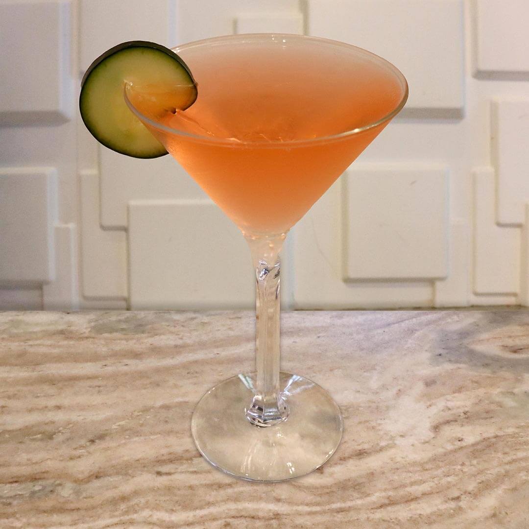 A light pinkish-red, translucent cocktail served in a chilled glass, garnished with a cucumber slice.