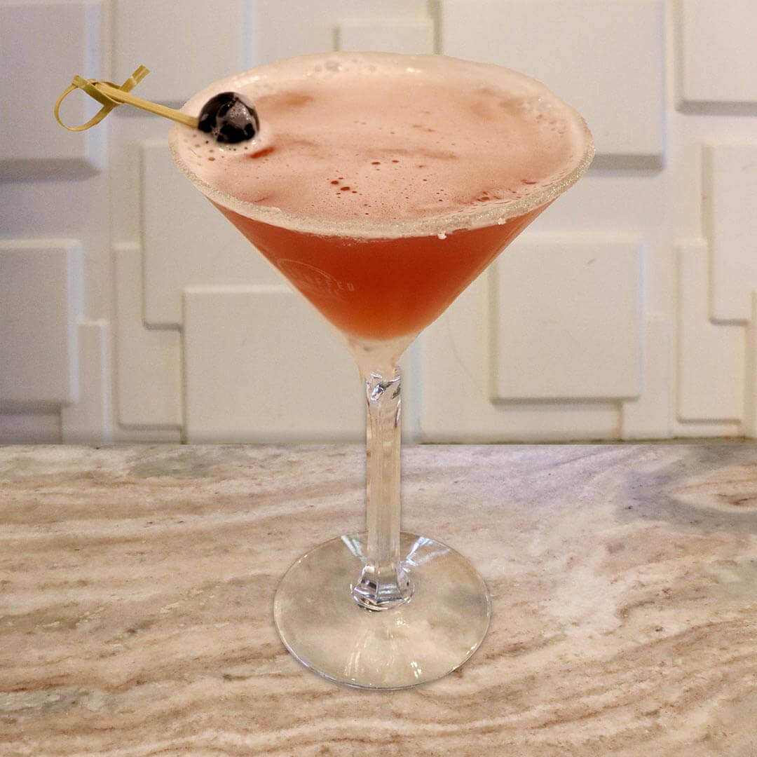 A bright crimson red cocktail with a sugared rim, garnished with a dark cherry.