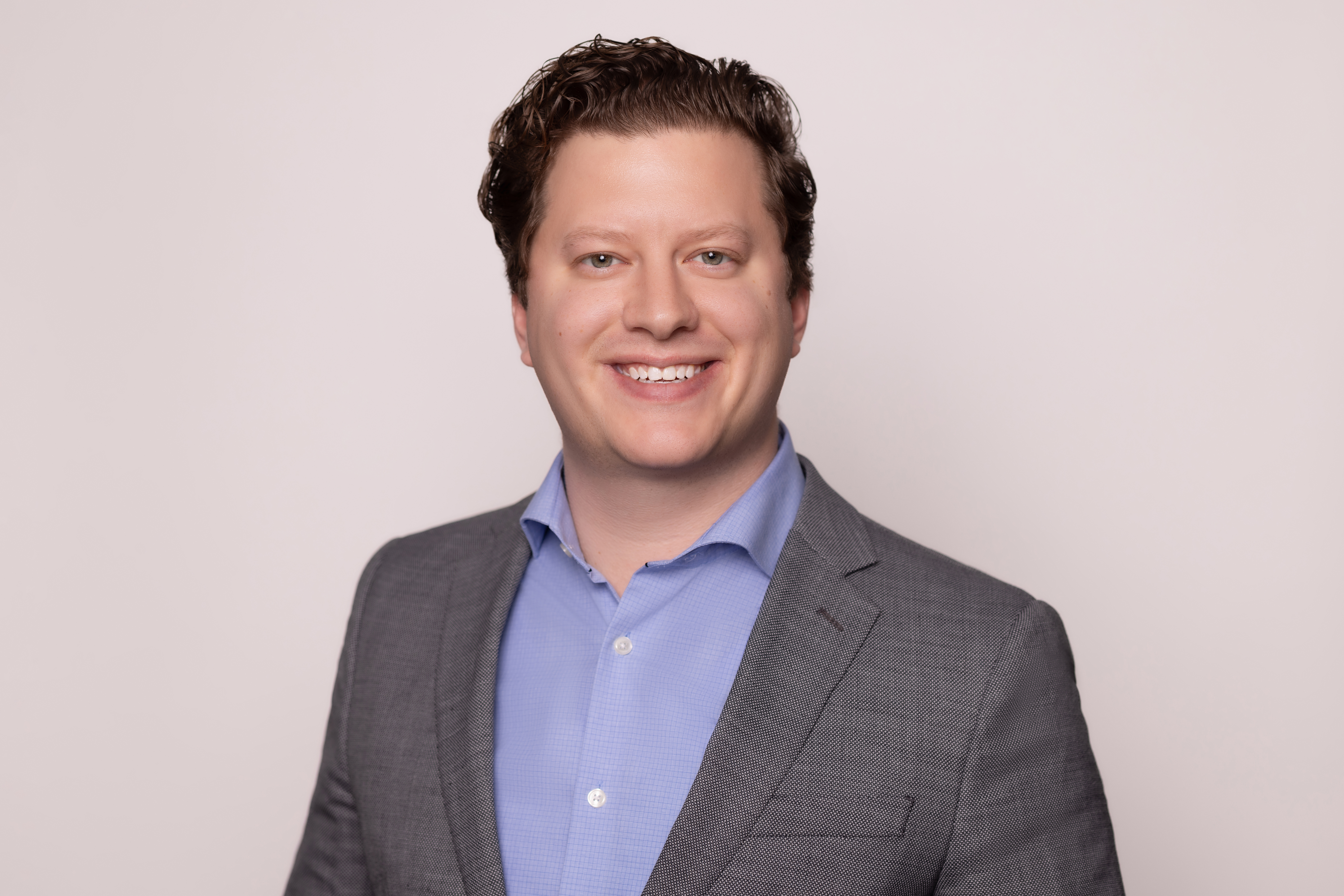 A man wearing a grey suit jacket and light-blue button-up dress shirt smiling at the camera.