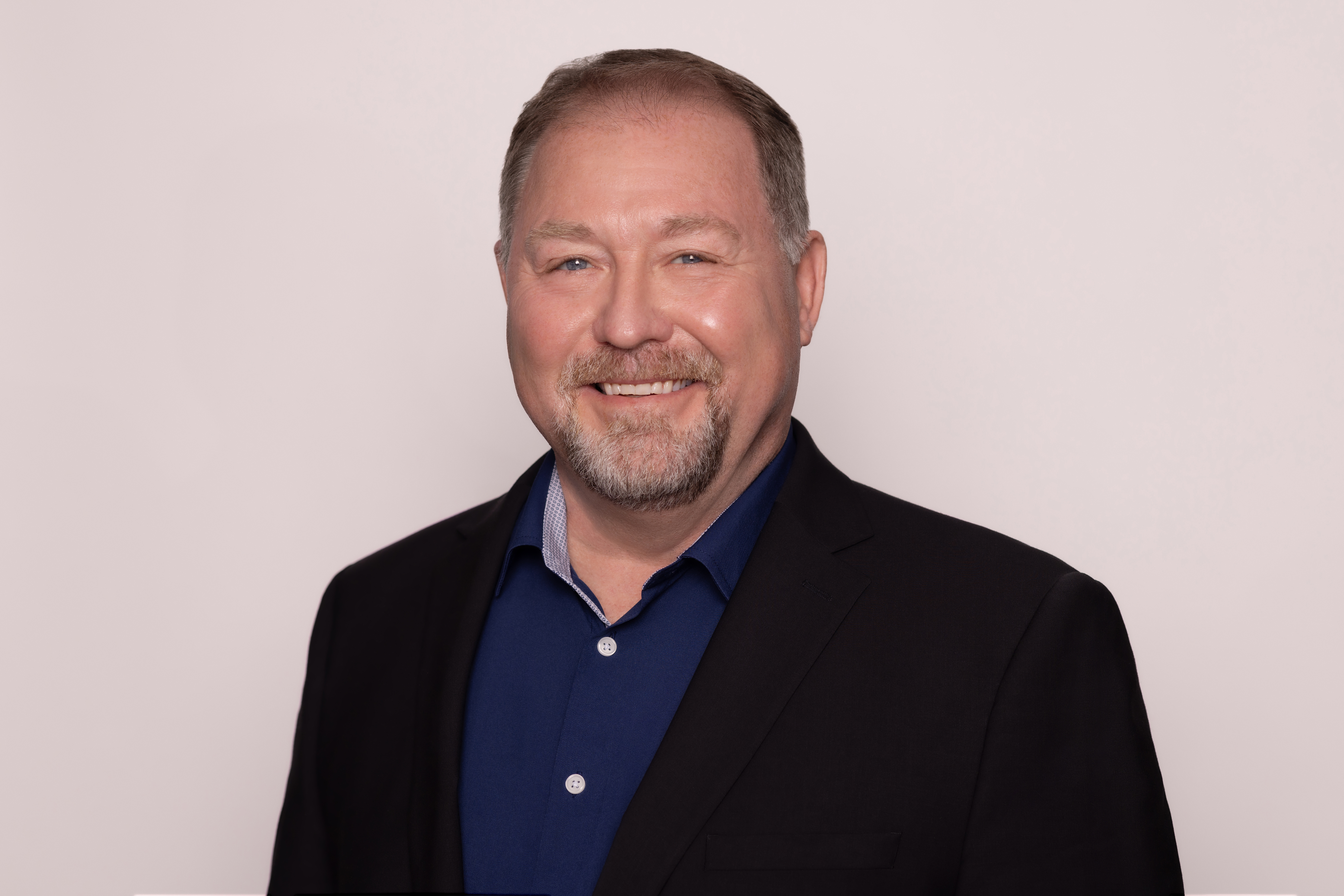 A man wearing a suit jacket and blue button-up dress shirt smiling at the camera.