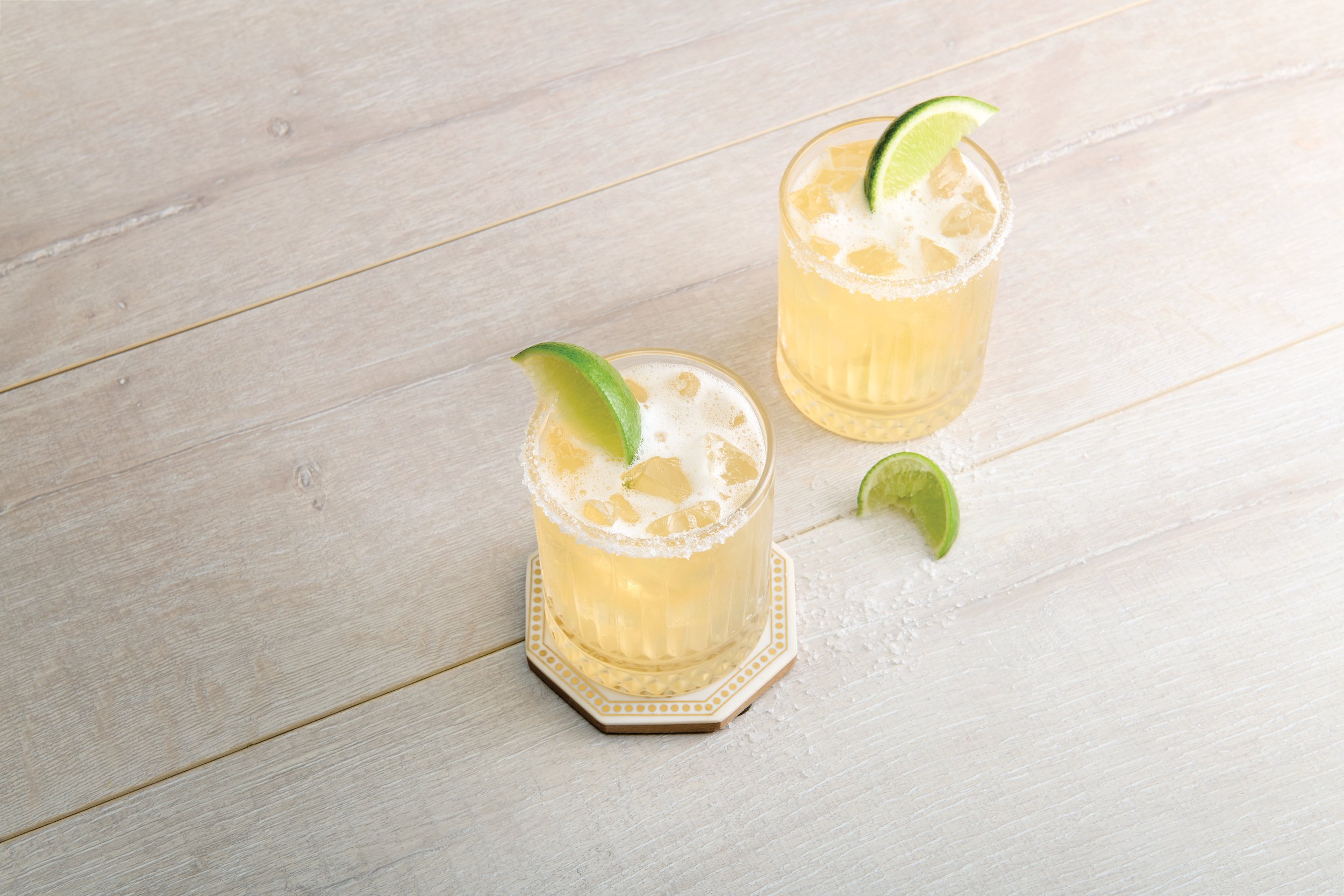 two glasses of yellow liquid with lime slices on a white surface