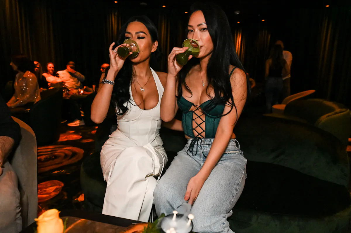 two women sitting on a couch drinking from a glass