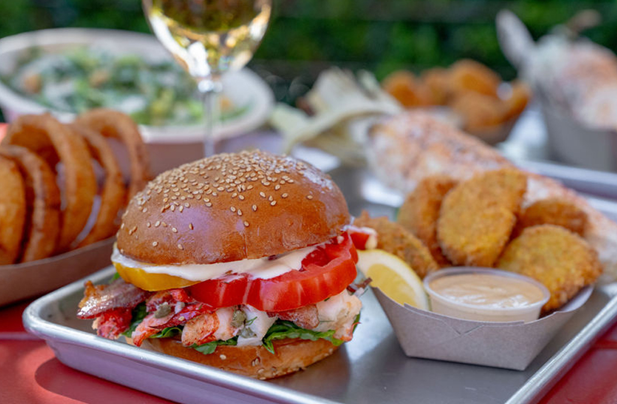 Lobster BLT with capers, herbs, and lemon mayo on a sesame bun with bacon, tomatoes, and little gem lettuce on a red picnic table with onion rings and fried green tomatoes.