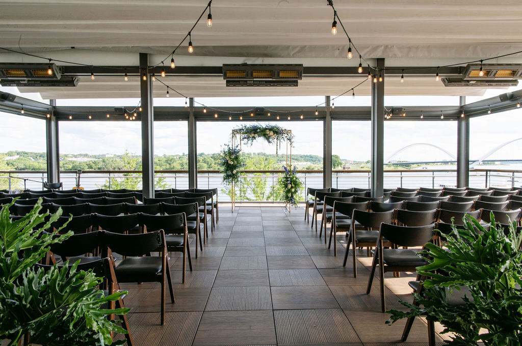 chairs and greenery are set up for a rooftop wedding at District Winery. at the center of the aisle, green ferns and plants hand from a geometric arch