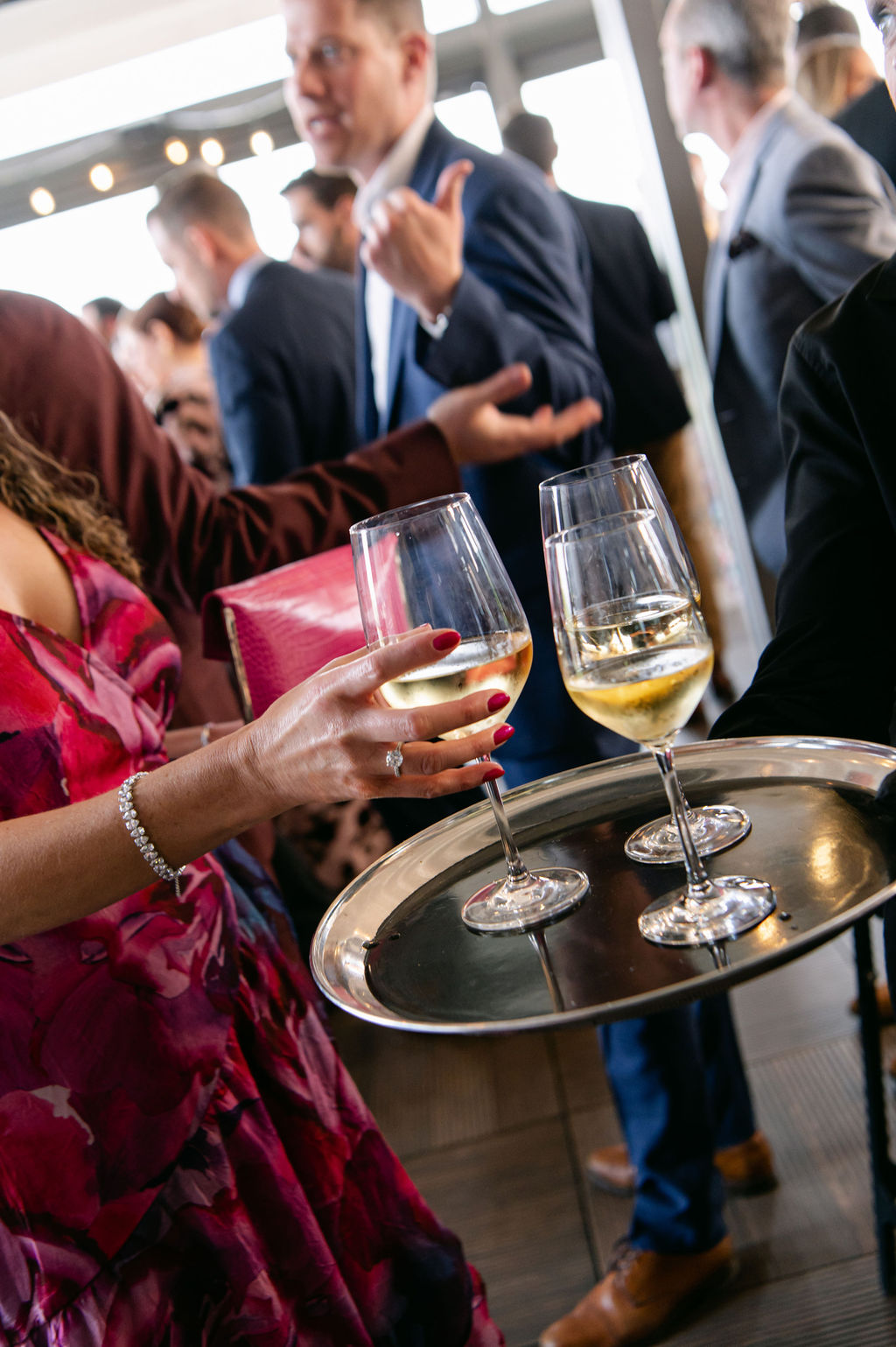 a woman in a pink dress reaches for a glass of District Winery wine wine being passed on a tray, behind her guests chat and smile