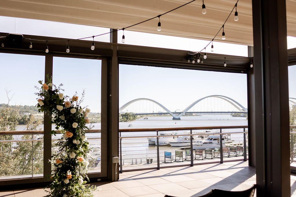 a close up of an asymmetrical wedding arch made from greenery and pink flowers. behind the archway you can see the Anacostia River and the Frederick Douglas memorial bridge