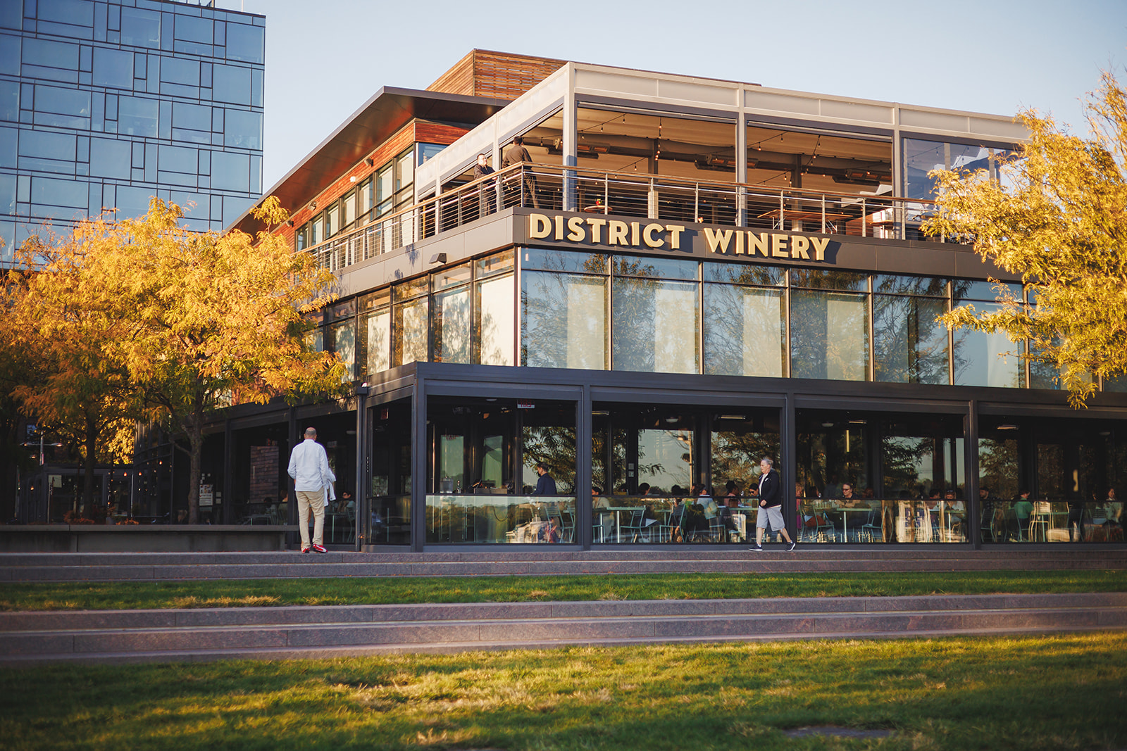 the exterior of district winery on a fall day. the rooftop is open and you can see tables set for an event. on the bottom floor diners enjoy a meal. there is yellow foliage on the trees.