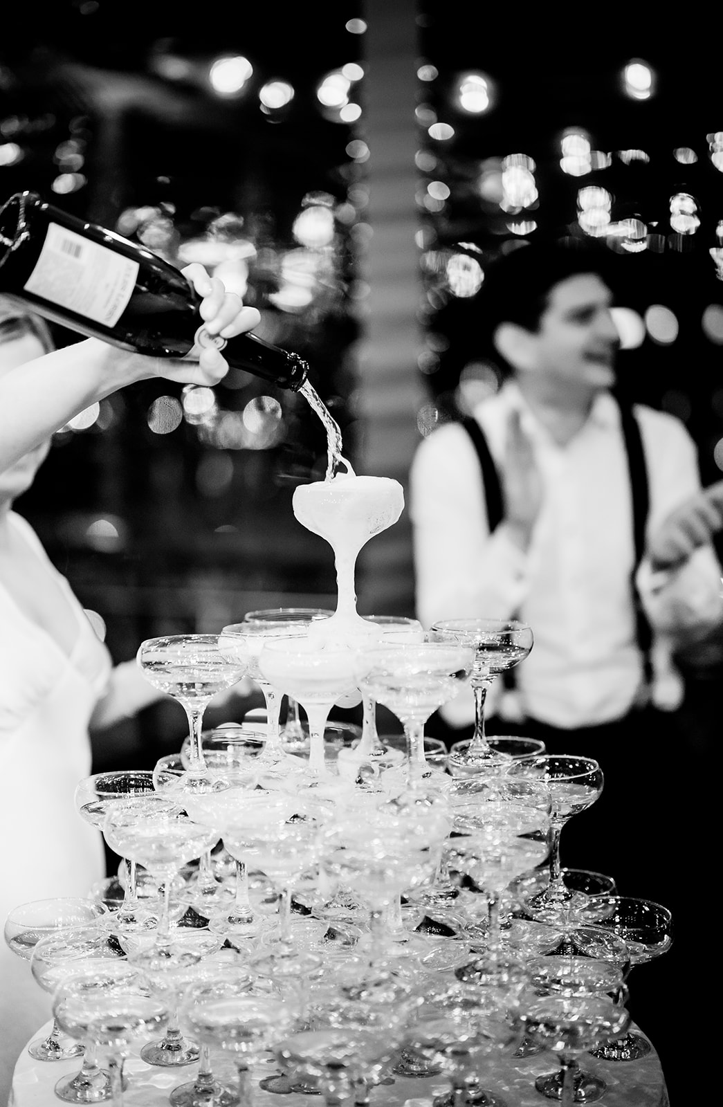 sparkling wine fizzes as a bride pours champagne into the top glass of a champagne tower