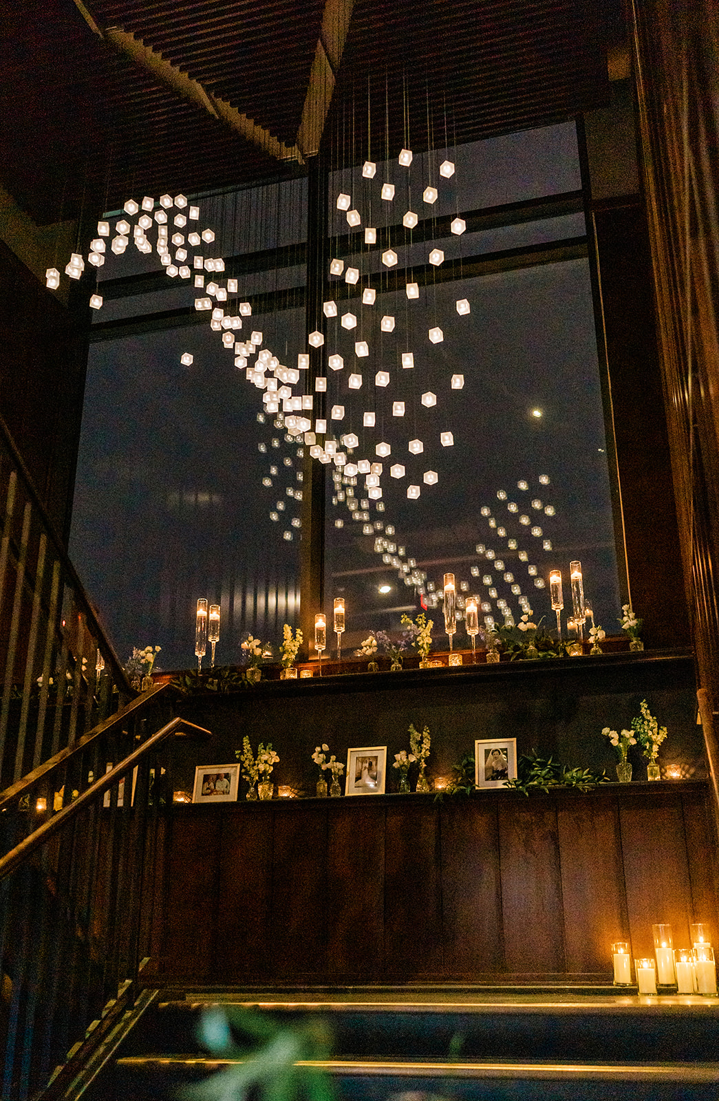 a chandelier hangs on a stairwell in low light. candles, flowers, and and photos line the wall as decor for a wedding