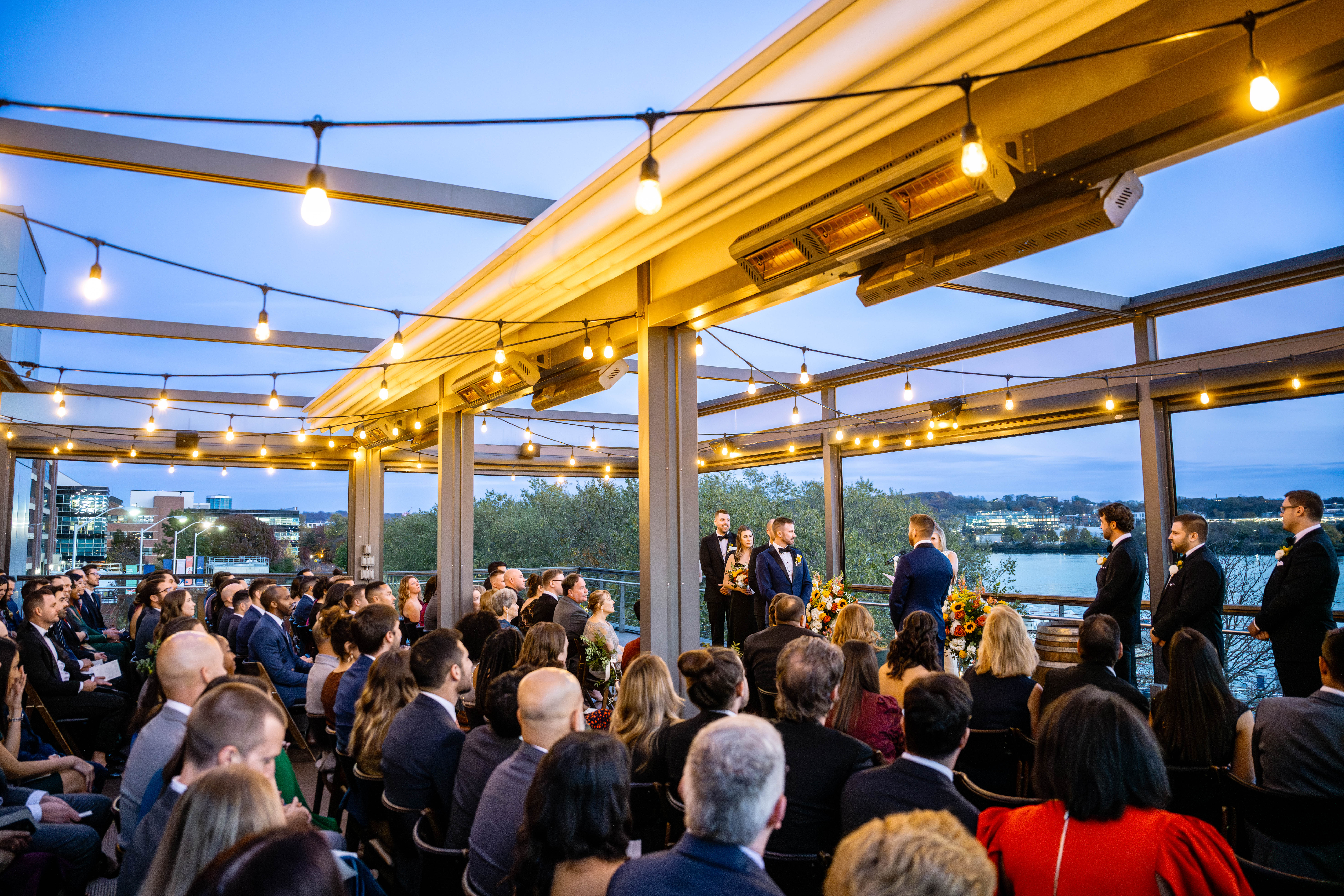 a crowd sits on the District Winery rooftop and watches a wedding ceremony as an LGBTQ couple exchanges vows. The rooftop overlooks the river and is lit with string lights