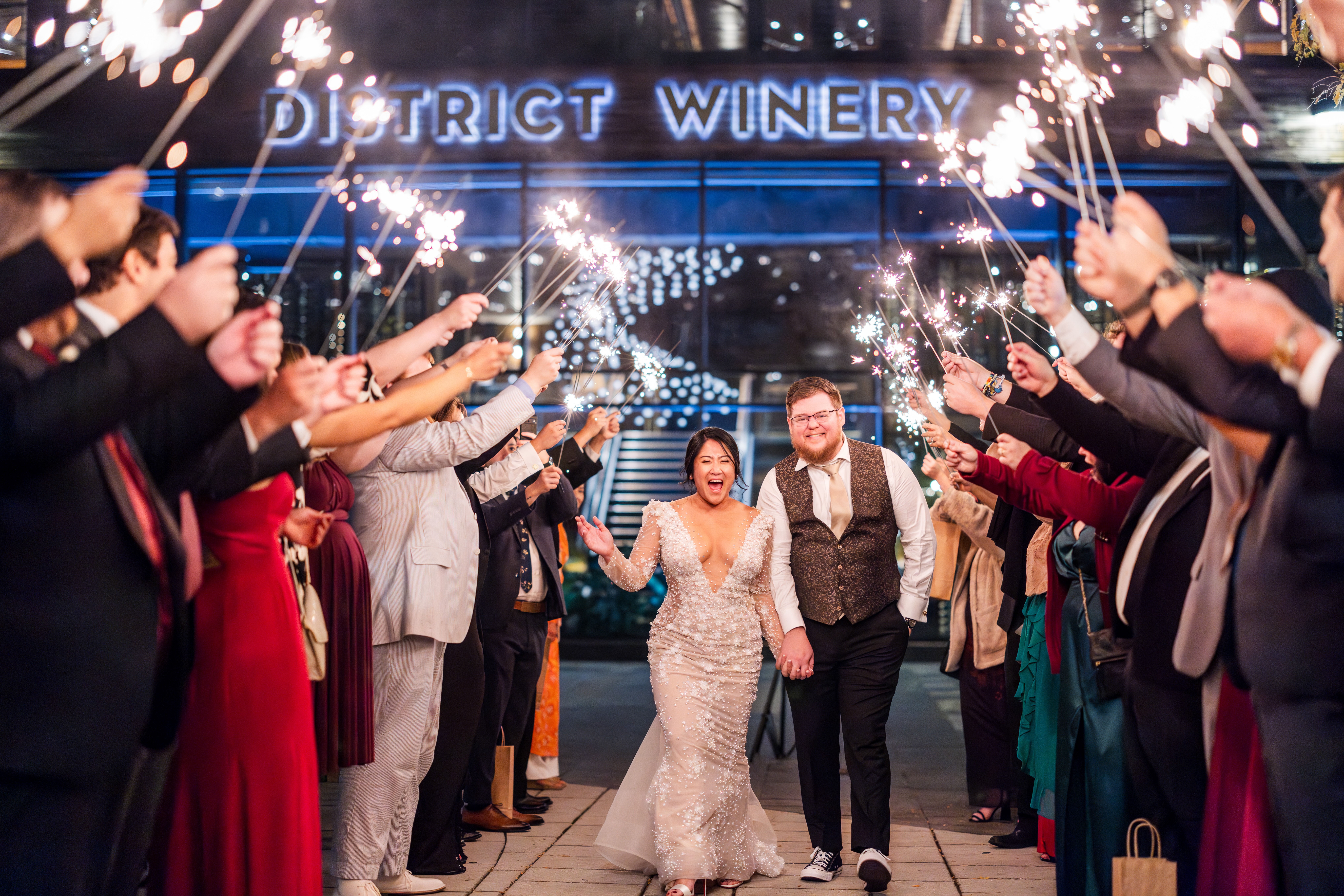 a wedding couple smiles as they walk through a row of sparklers held up by their guests