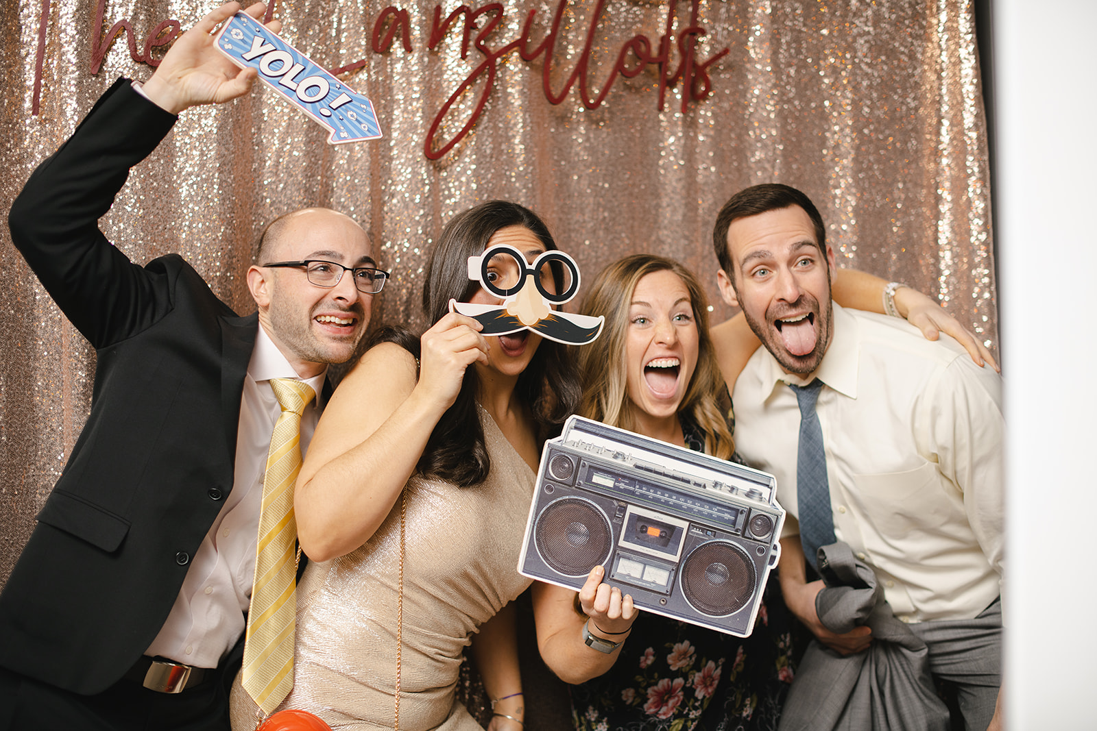 four wedding guests laugh and make faces in a photo booth. they hold a fake stereo and mustache as props