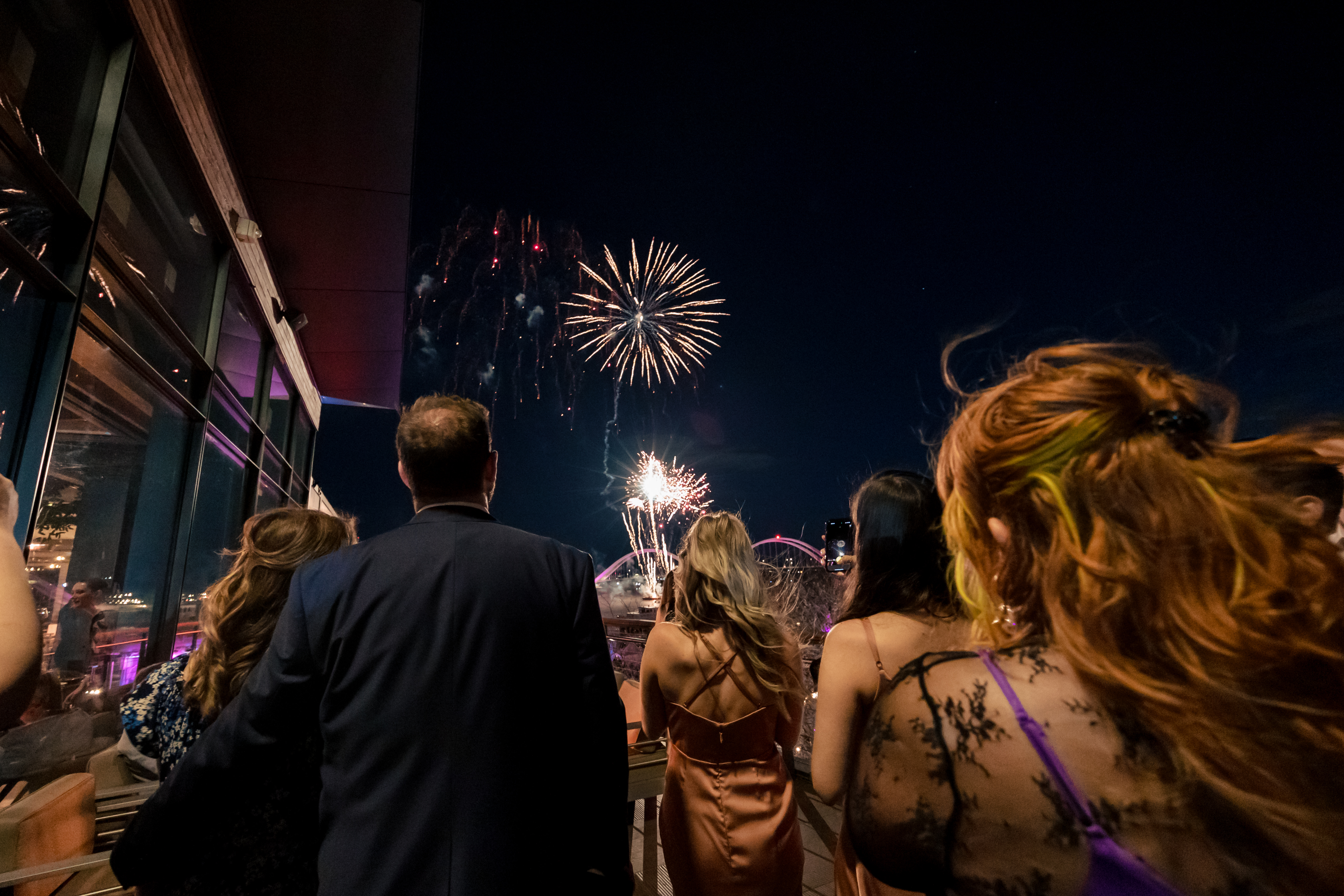 a group of event guests have their backs to the camera as they look up at fireworks