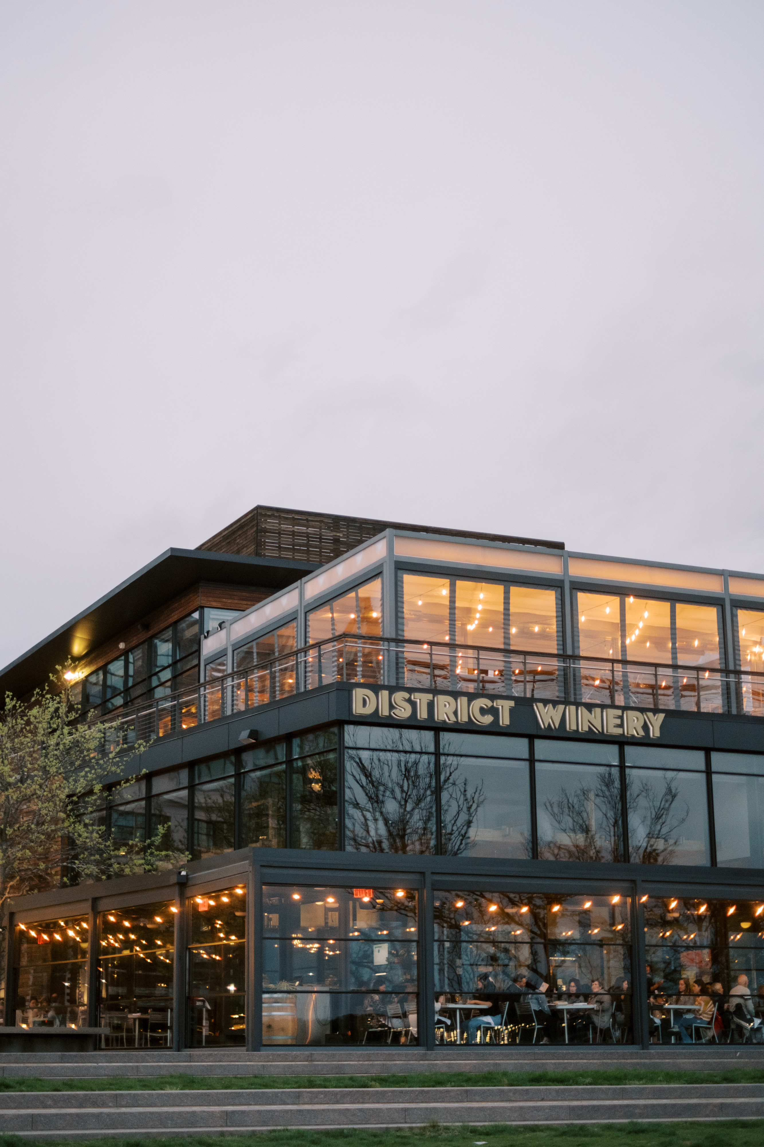 the exterior of District Winery at twilight. You can see people dining and mingling inside. Both floors are warm and lit with string lights