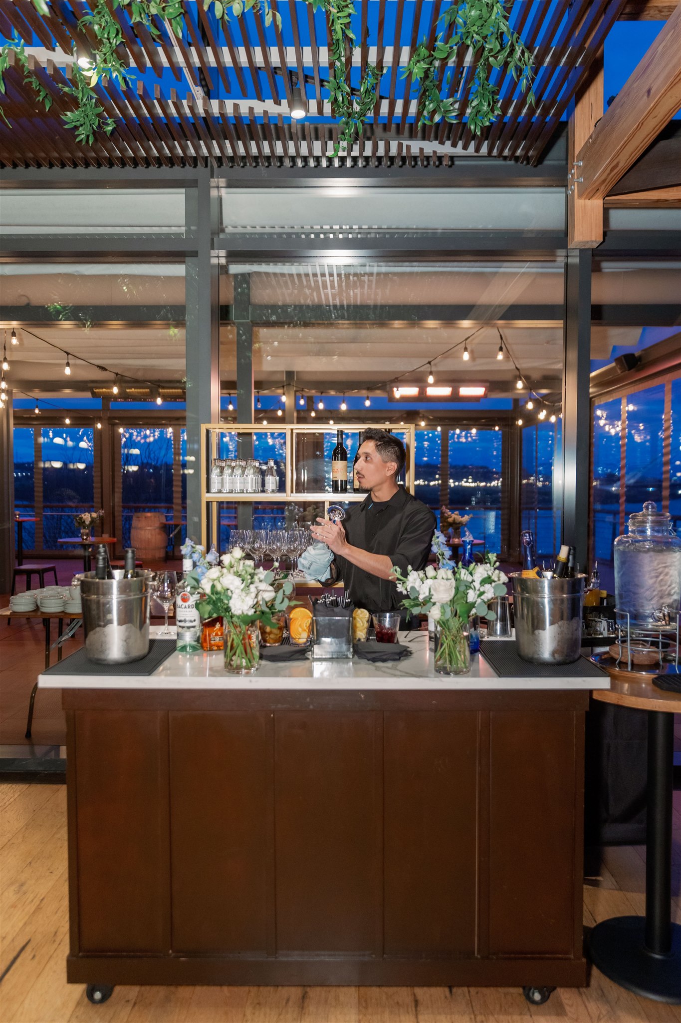 a bartender shakes a drink at a white marble bar. behind him the sun is setting over the anacostia river and the space is warm with fairy lights