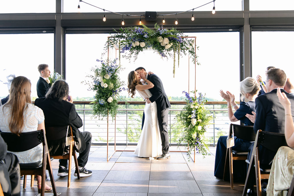 a bride and groom share their first kiss at the alter in front of a square geometric modern wedding arch covered in flowers. guests look on and clap