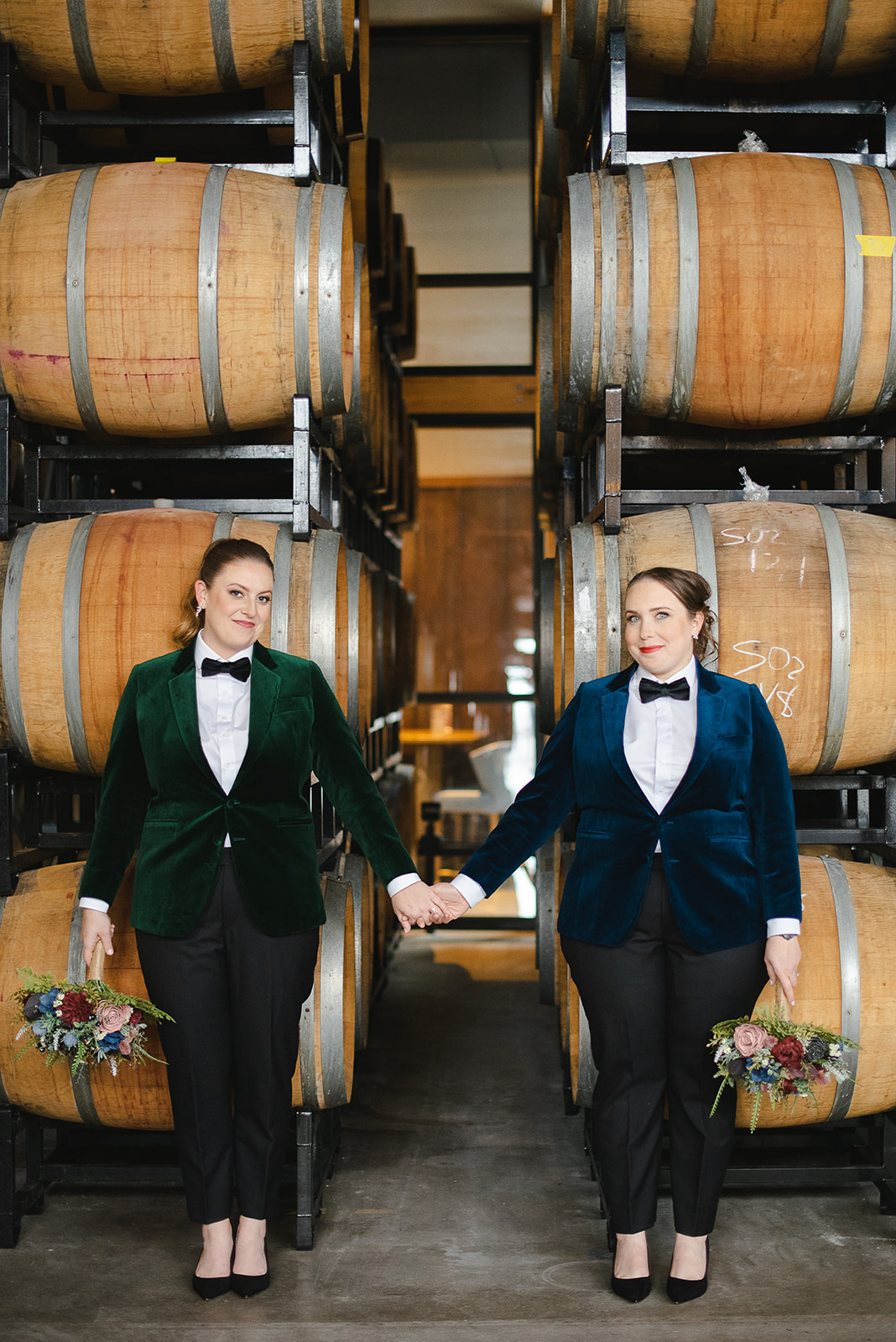 a couple poses in front of wine barrels holding a wedding bouquet, the bride on the left is wearing an emerald suite and the bride on the right is wearing a navy suite