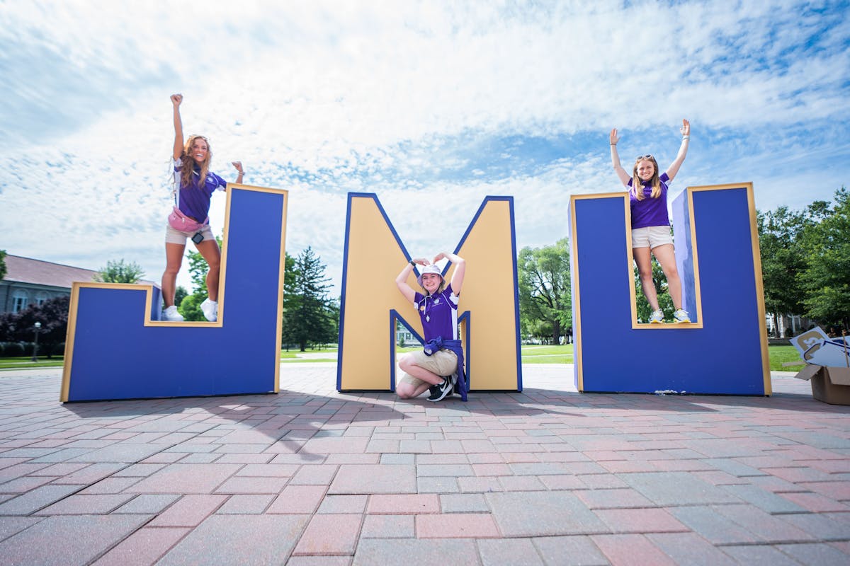 JMU letters on the Quad in front of Wilson Hall with students posing and smiling