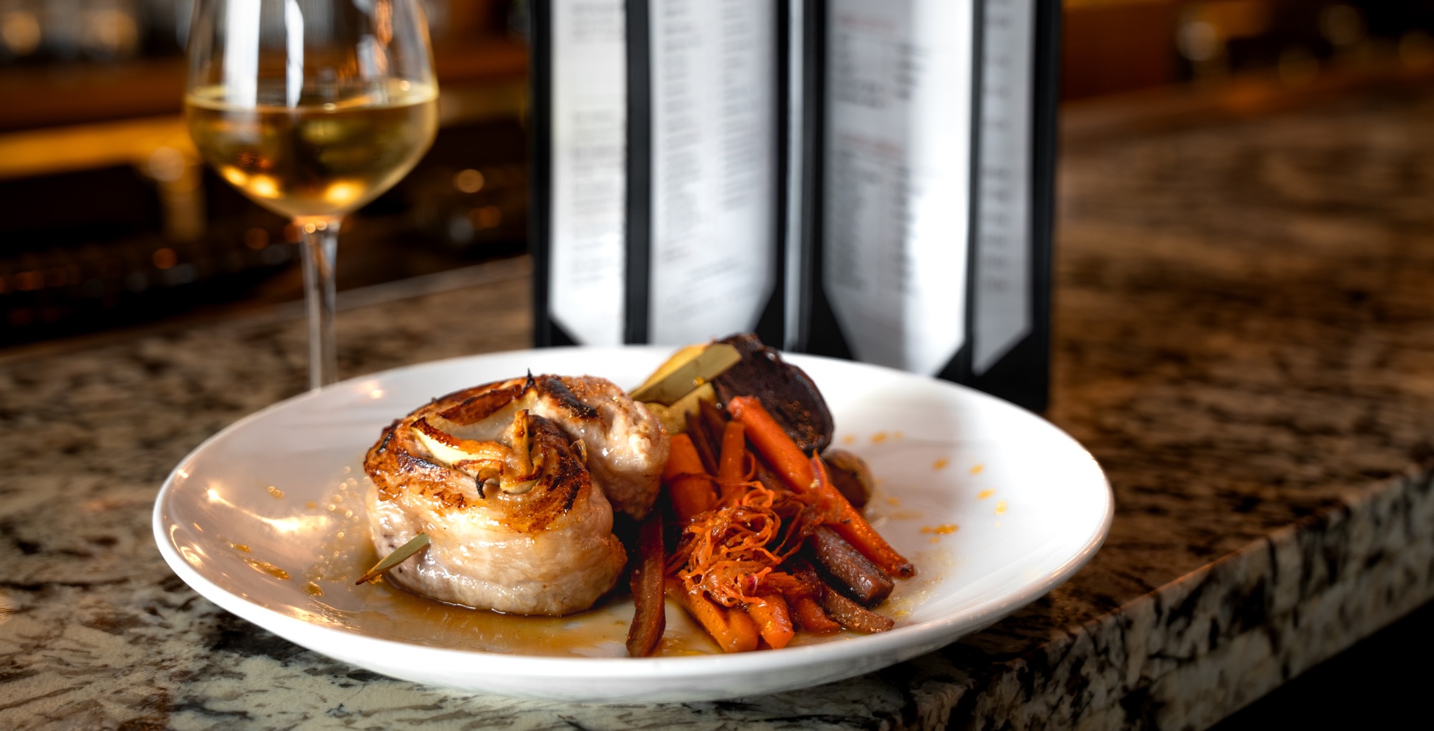 stuffed pork chop entree with glass of white wine and menu on bar top