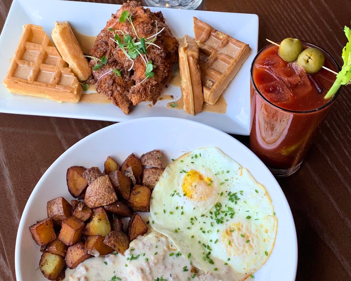 brunch plates with chicken and waffles, biscuits and gravy with eggs and potatoes, and a bloody mary cocktail