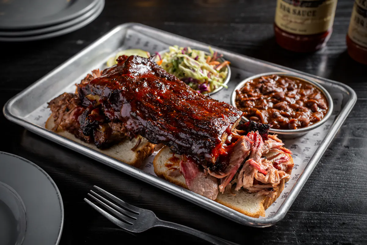 A tray of BBQ ribs and pulled pork on bread with coleslaw and baked beans.