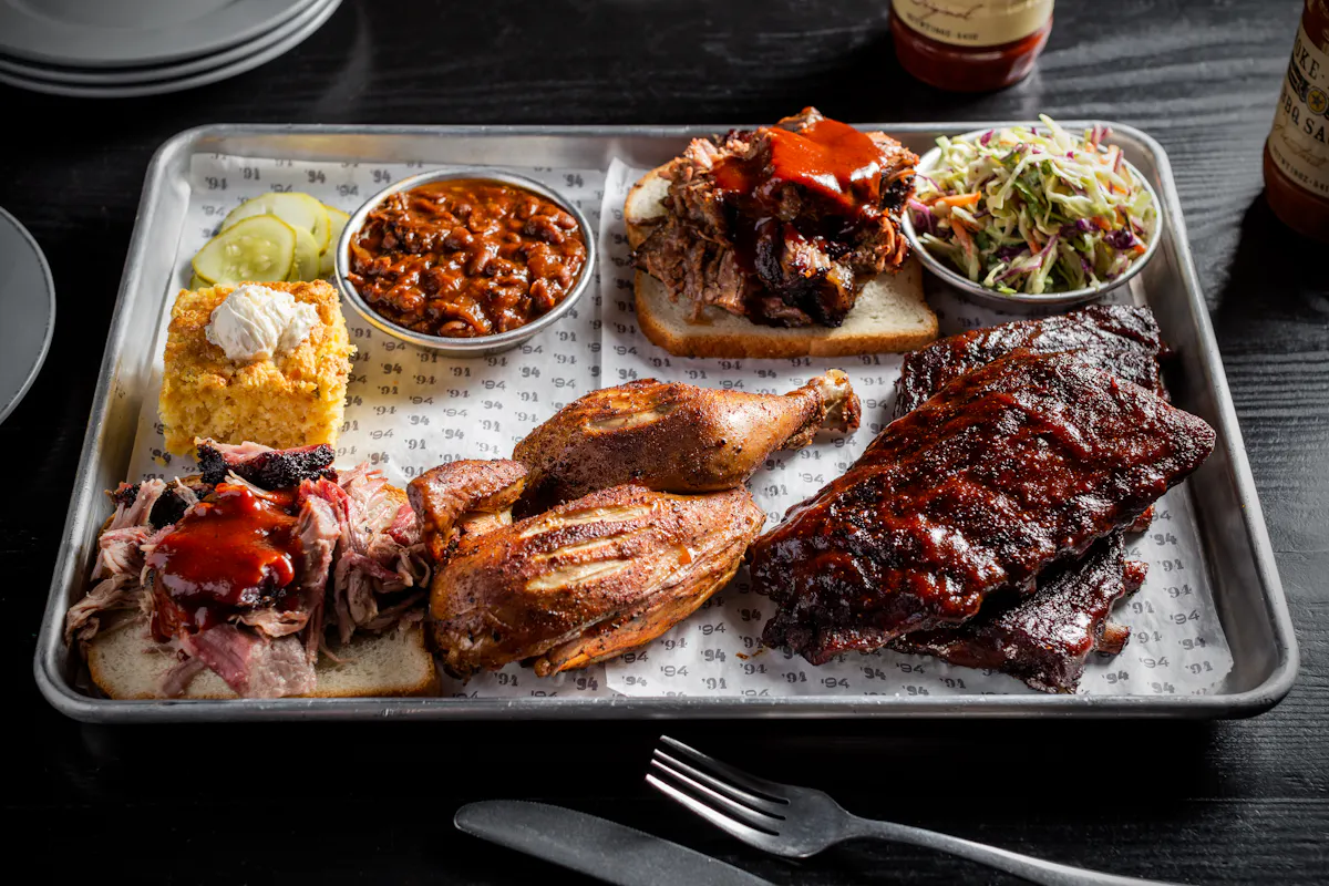 A large platter featuring a variety of BBQ meats including ribs, chicken, pulled pork, and brisket, with sides of cornbread, beans, and coleslaw.