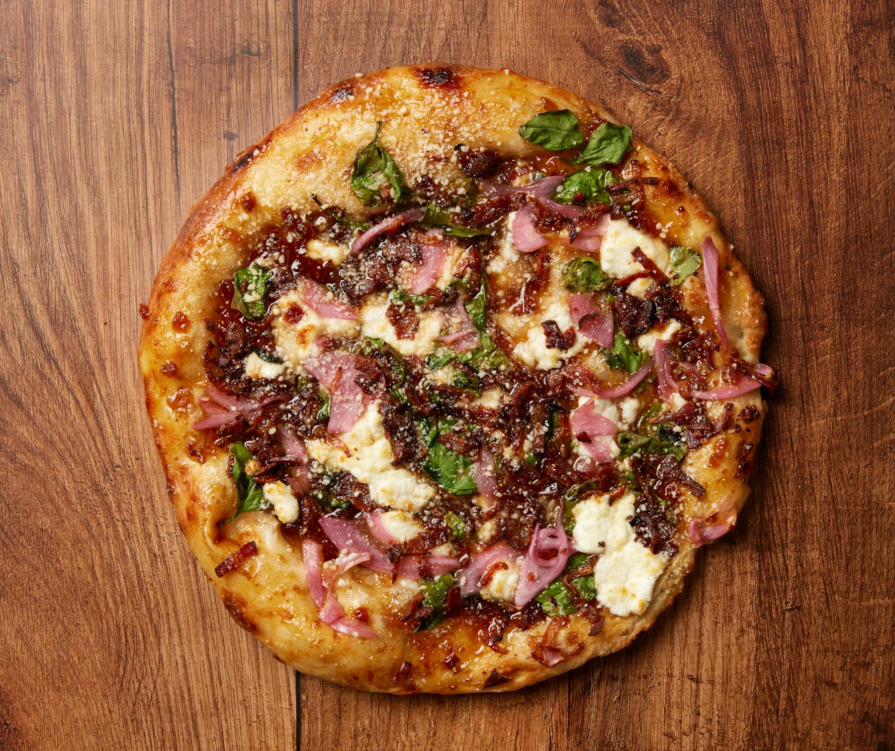 A circular cooked pizza with figs, prosciutto, and goat cheese on a rustic wooden surface.