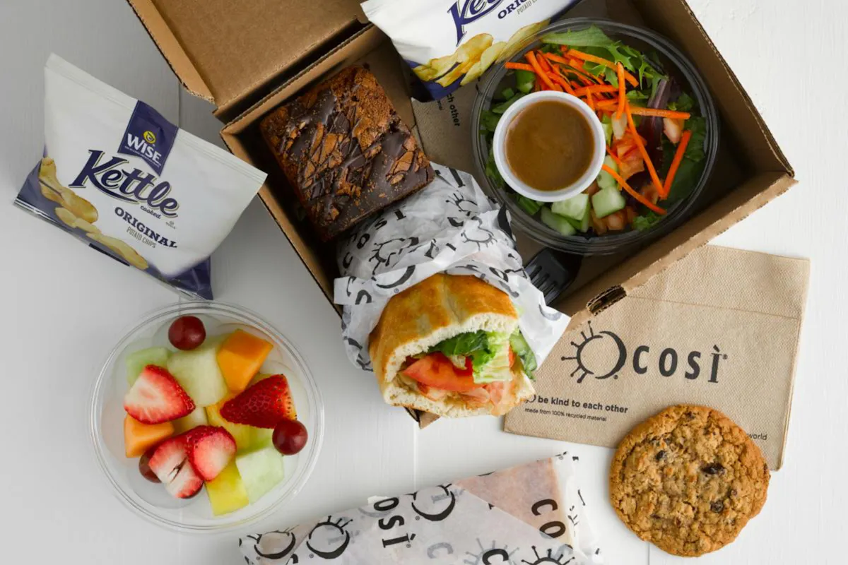 A Cos&igrave; boxed lunch with a wrapped sandwich, side salad, fruit cup, chips, a brownie, and a cookie
