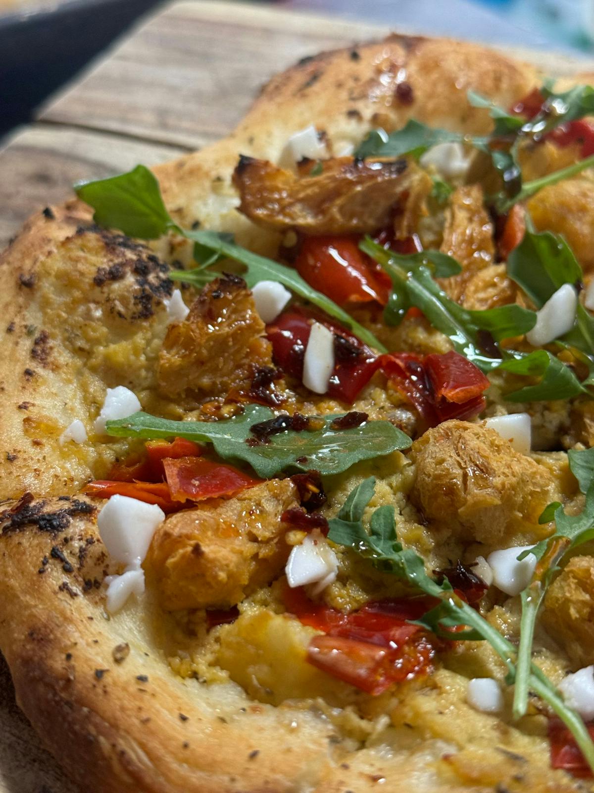 A pizza featuring chicken, cheese, and greens is artfully arranged on a wooden cutting board, ready to be enjoyed