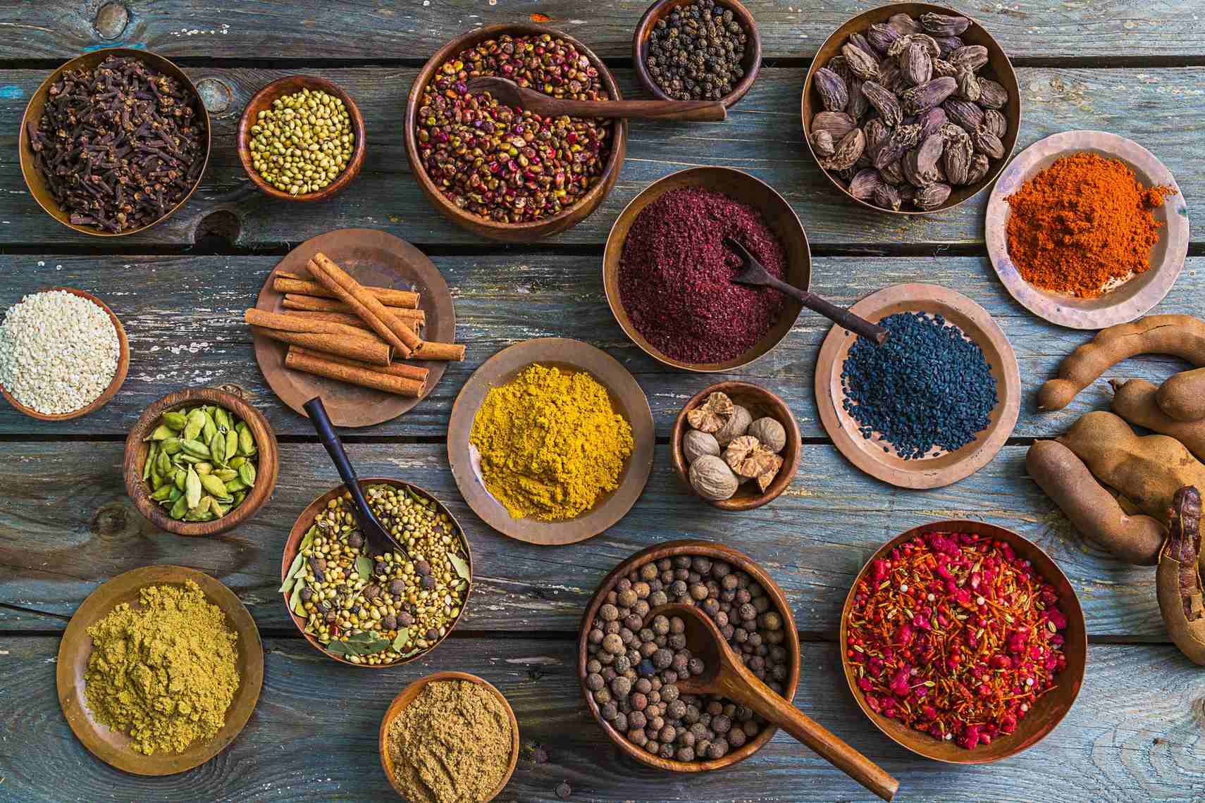 An array of spices and seeds in wooden bowls on a rustic wooden surface.