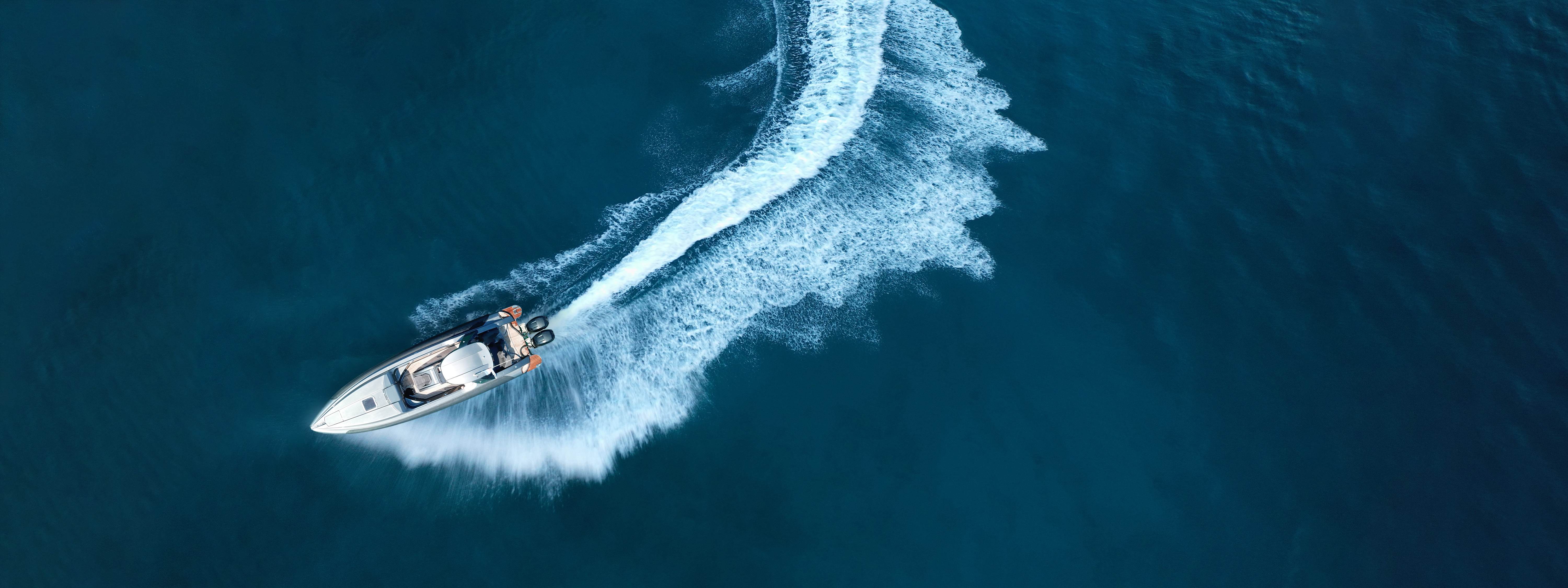 A sleek luxury speedboat cuts through deep blue ocean water, leaving a dynamic white wake behind it. Captured from an aerial perspective, the boat curves gracefully across the water, highlighting motion and elegance against the vast, serene sea.