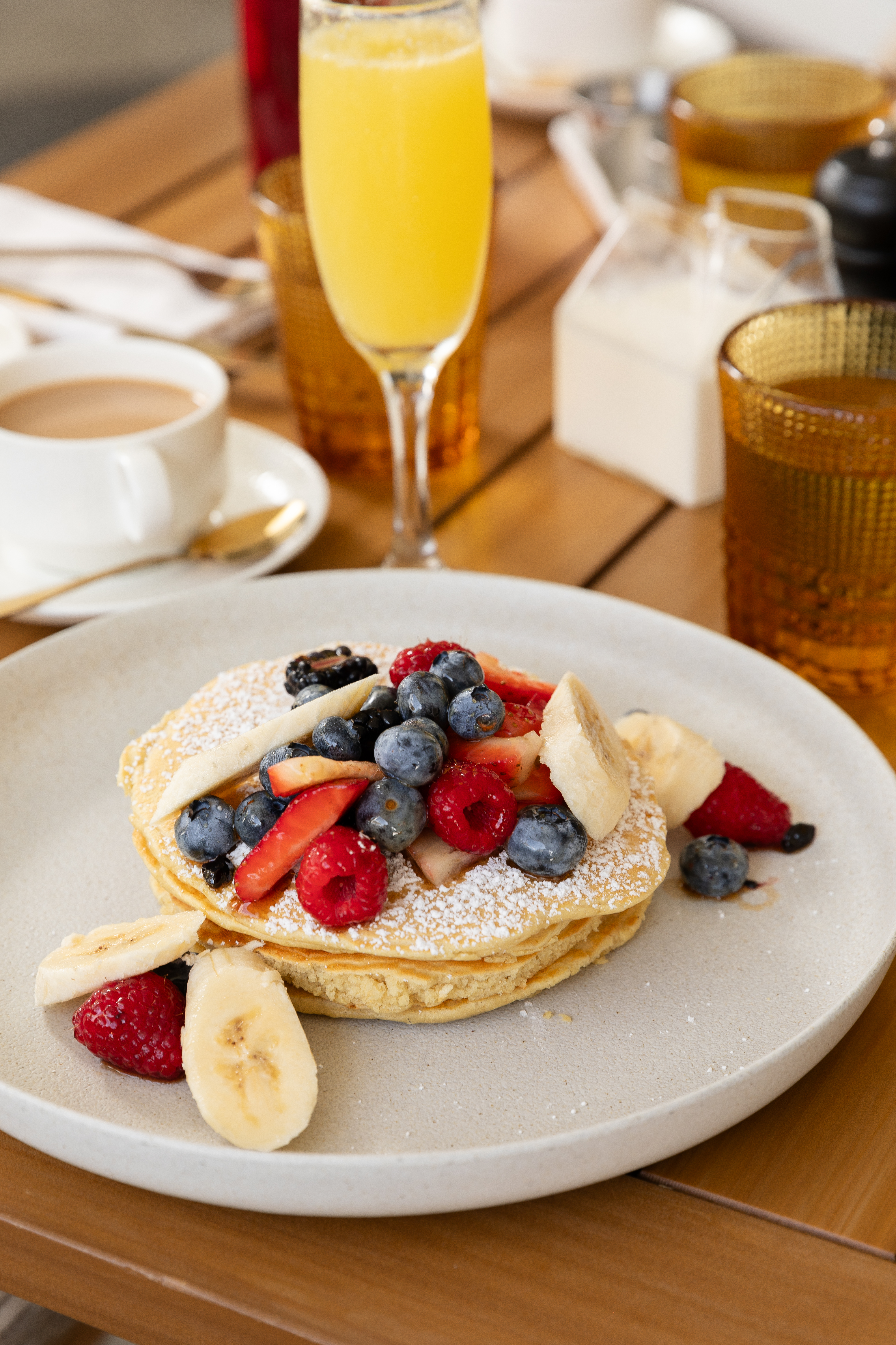 A plate of fluffy pancakes topped with powdered sugar, fresh blueberries, raspberries, strawberries, and banana slices sits on a wooden table. Surrounding the dish are a glass of orange mimosa, a cup of coffee with a gold spoon, amber drinking glasses, and a small milk pitcher, creating a warm and inviting breakfast setting.