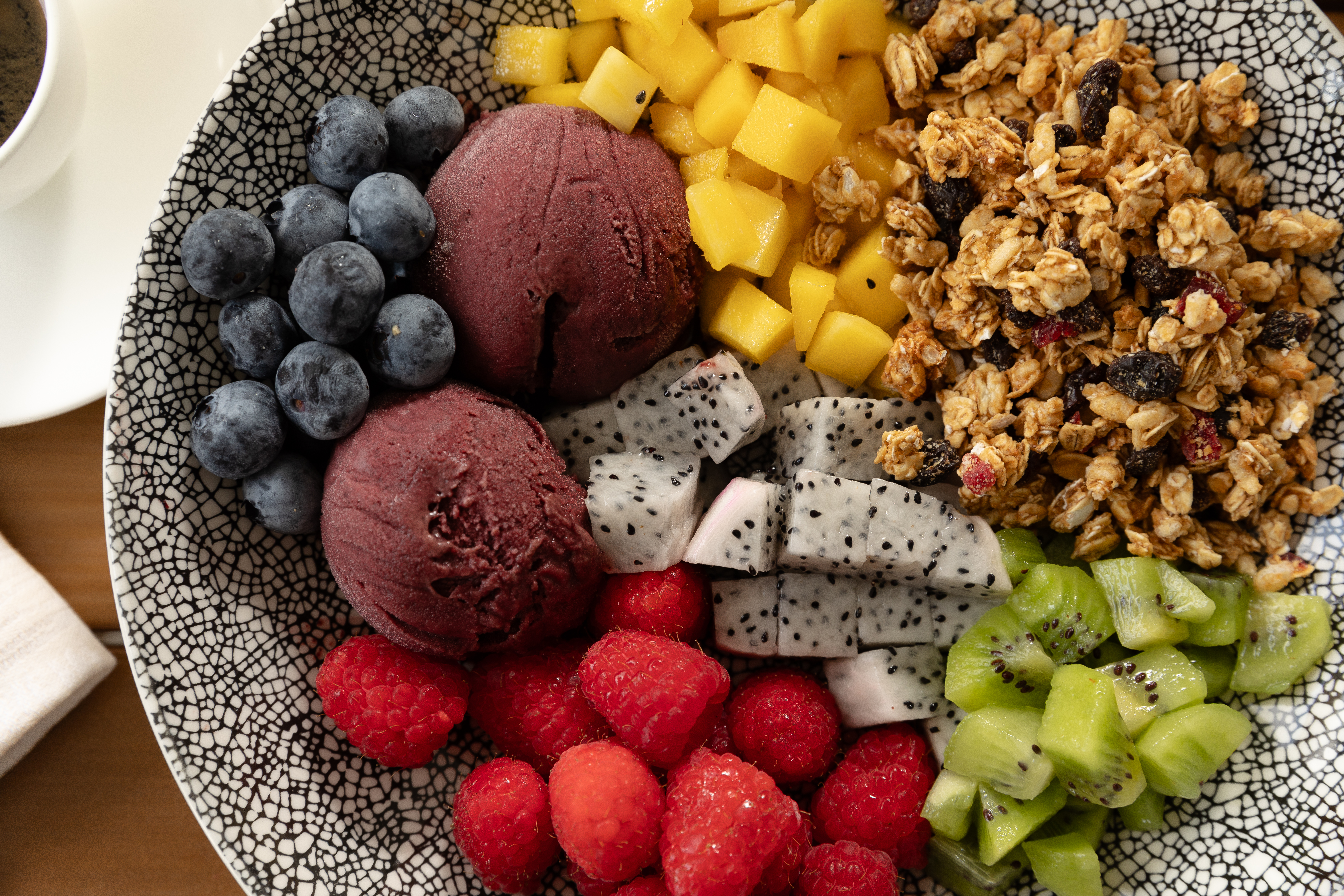 A vibrant acai bowl topped with fresh fruit and granola, served in a patterned bowl. The toppings include blueberries, raspberries, kiwi, diced mango, cubed dragon fruit, crunchy granola with raisins, and two scoops of deep purple acai. The colorful ingredients are neatly arranged, creating a visually appealing and healthy breakfast dish.