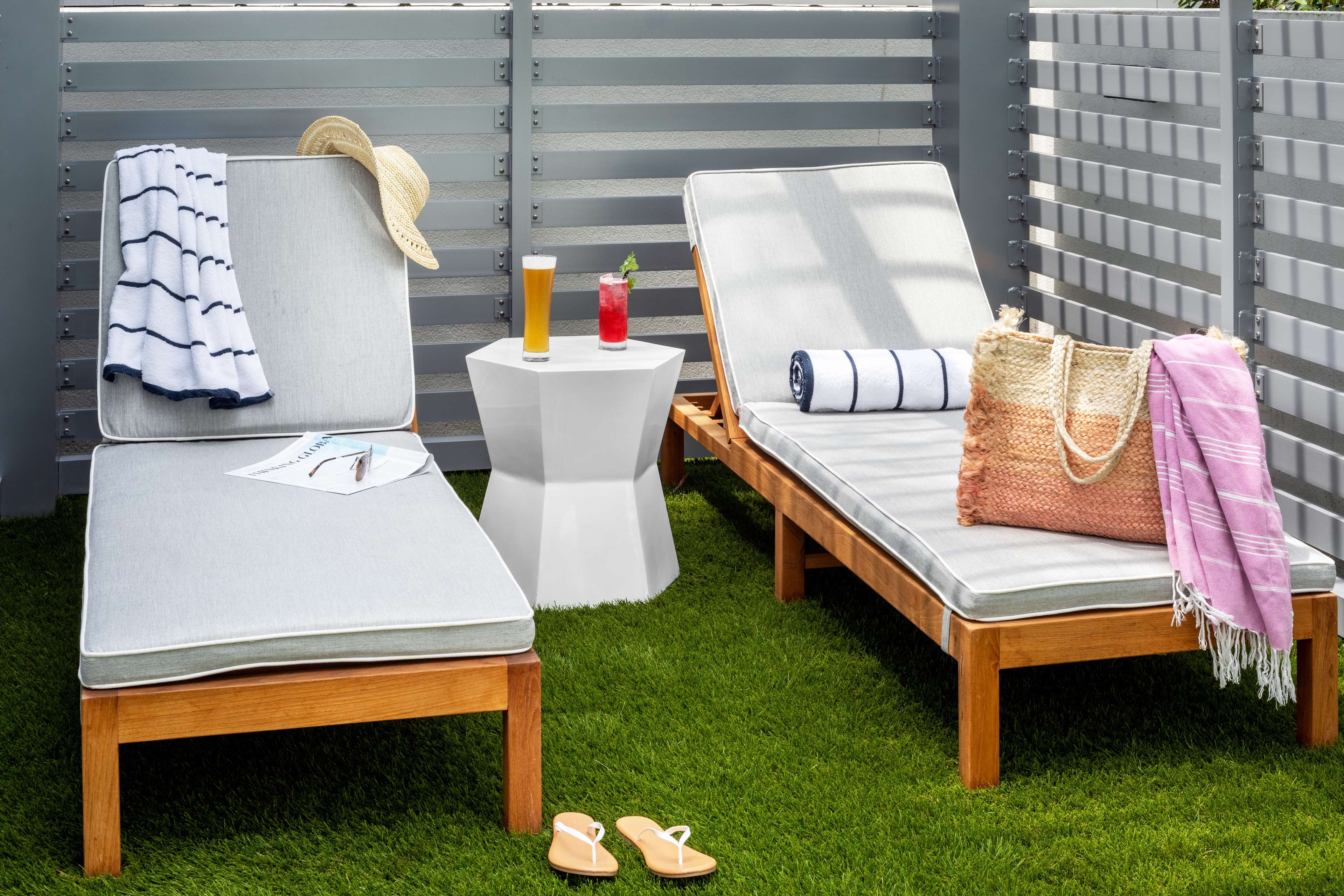 Two wooden lounge chairs with light gray cushions are set on green artificial grass beside a modern white side table. One chair has a sunhat, striped towel, sunglasses, and a magazine, while the other holds a rolled towel, a straw beach bag, and a pink Turkish towel. On the table between them are two refreshing drinks—a beer and a red cocktail. A pair of beige flip-flops sits on the grass, and the area is enclosed by sleek gray slatted fencing, creating a cozy private cabana vibe.
