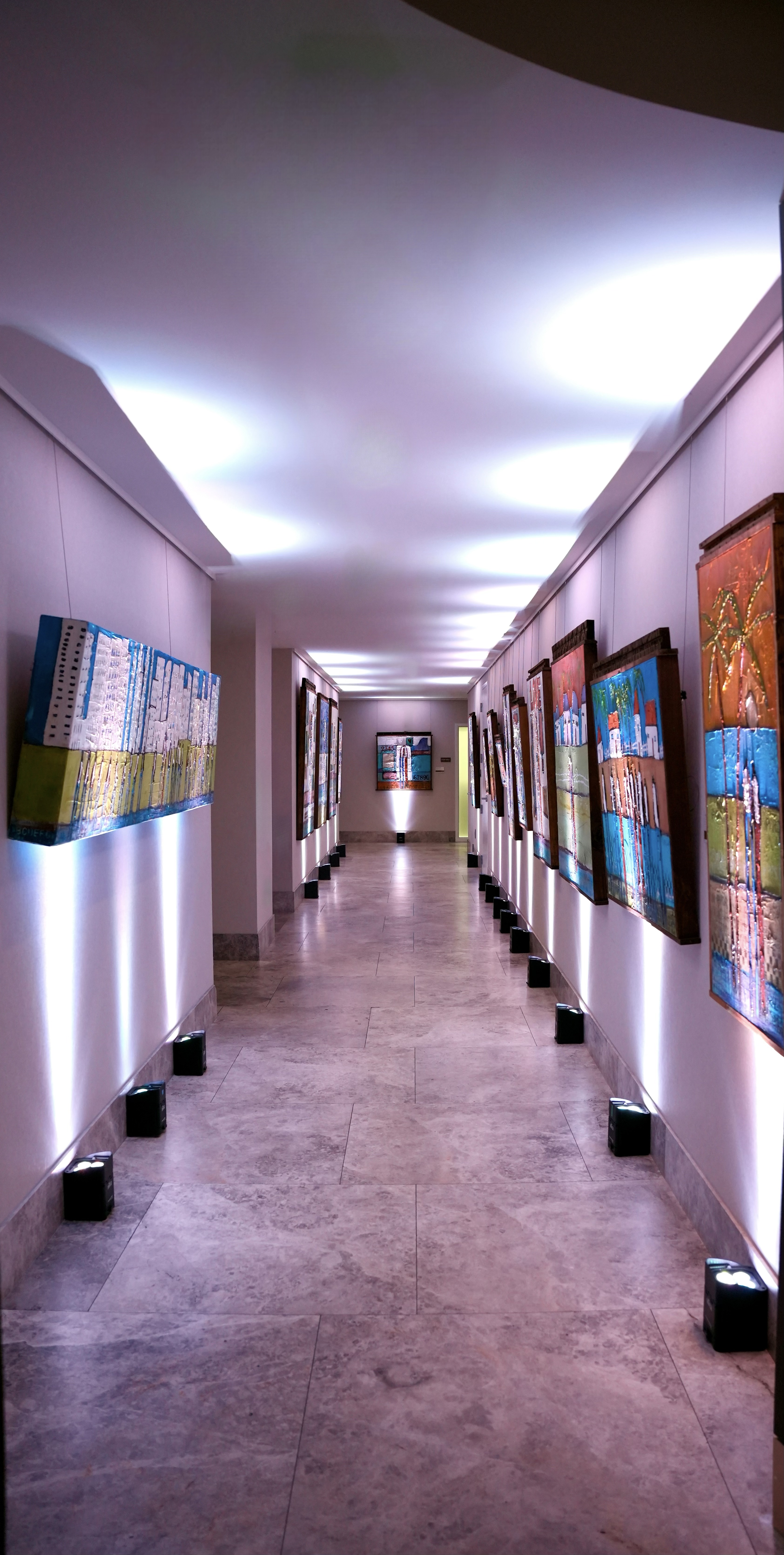 A modern hallway with polished stone flooring and bright overhead lighting features colorful paintings displayed on both walls. Small floor lights highlight each artwork, creating a gallery-like atmosphere. A larger painting is visible at the far end of the hallway, drawing attention down the corridor.