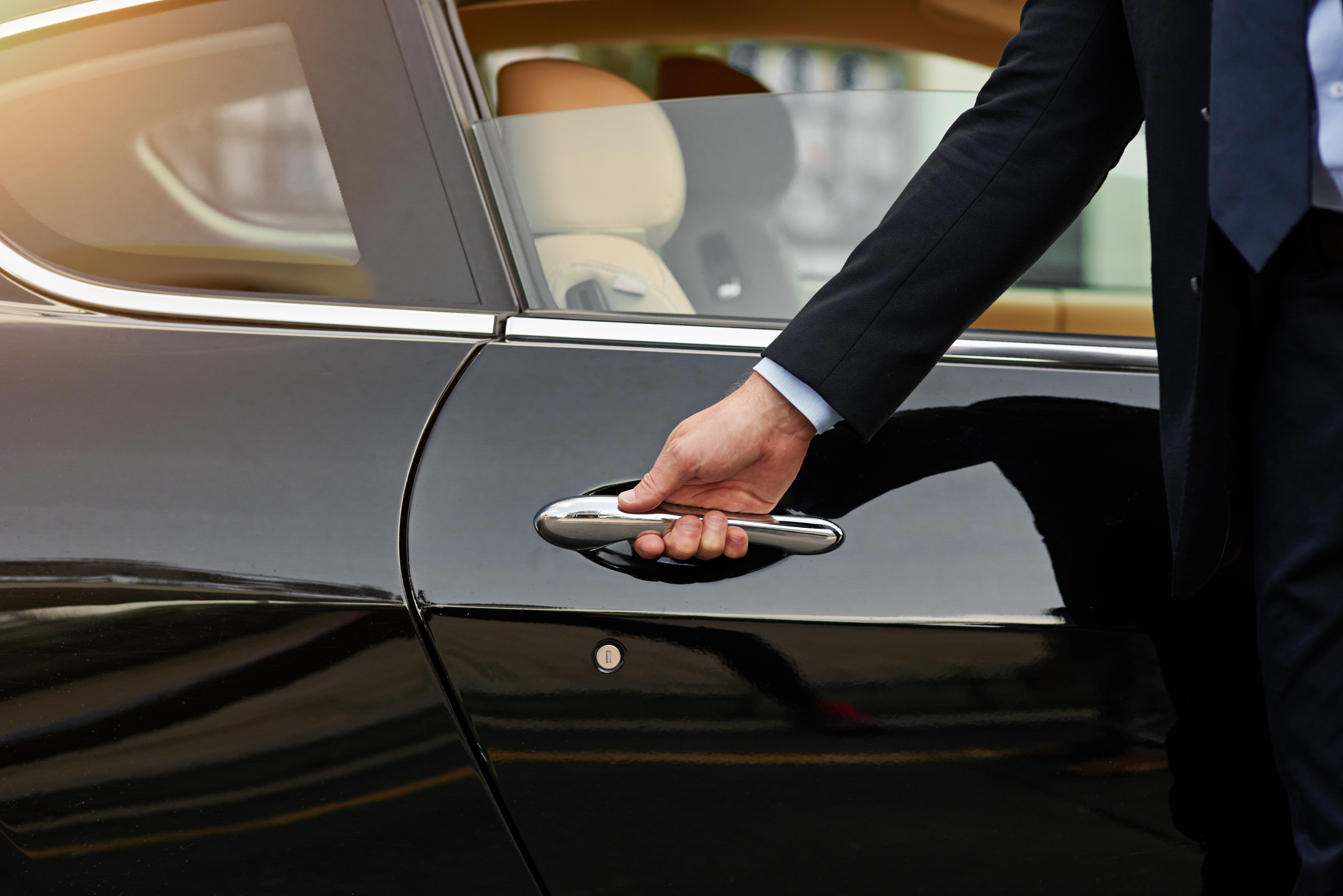 A professionally dressed man in a dark suit opens the door of a sleek black luxury car. The vehicle features a polished chrome handle and beige leather interior, suggesting a premium or VIP transportation service. The scene conveys elegance, service, and sophistication.