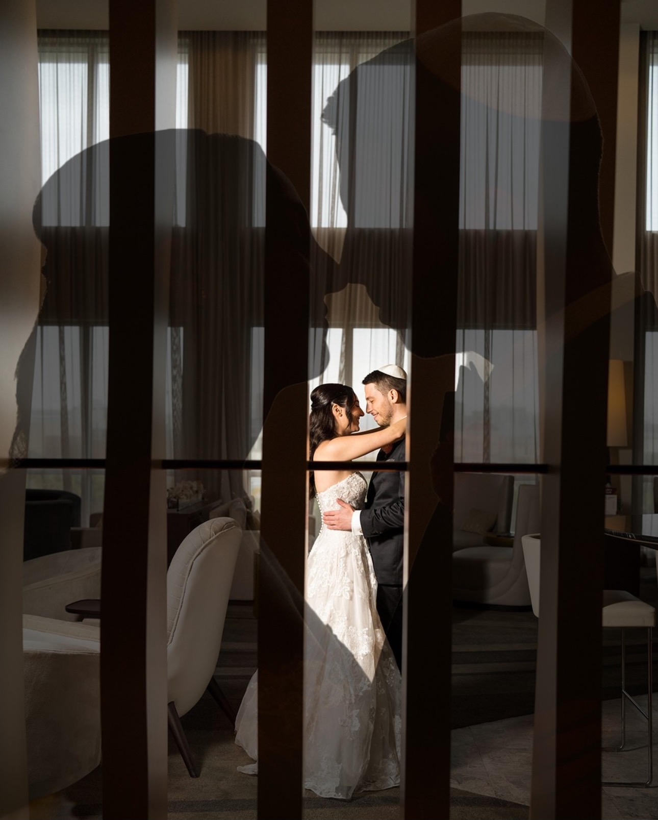A romantic and artistic photograph of a bride and groom embracing indoors, framed creatively through silhouetted profiles of their own faces. The couple stands in a softly lit, elegant room with floor-to-ceiling windows and sheer curtains. The bride wears a lace wedding gown, and the groom is in a formal suit with a kippah, creating a heartfelt and visually layered wedding portrait.